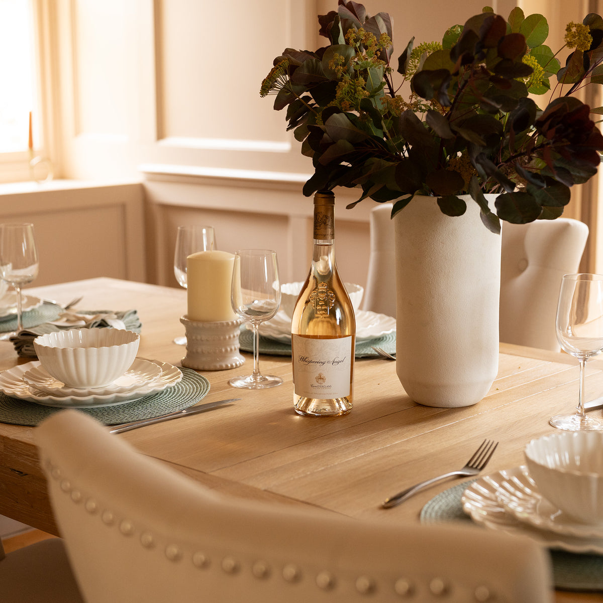 The Burford 8 Seat Extending Dining Set with Regent Chairs in Natural Oak and Ivory is beautifully arranged with wine, white dishes, candles, and a large vase of dark leafy flowers. This dining set features an extendable natural oak table, making it perfect for both intimate dinners and grand gatherings.