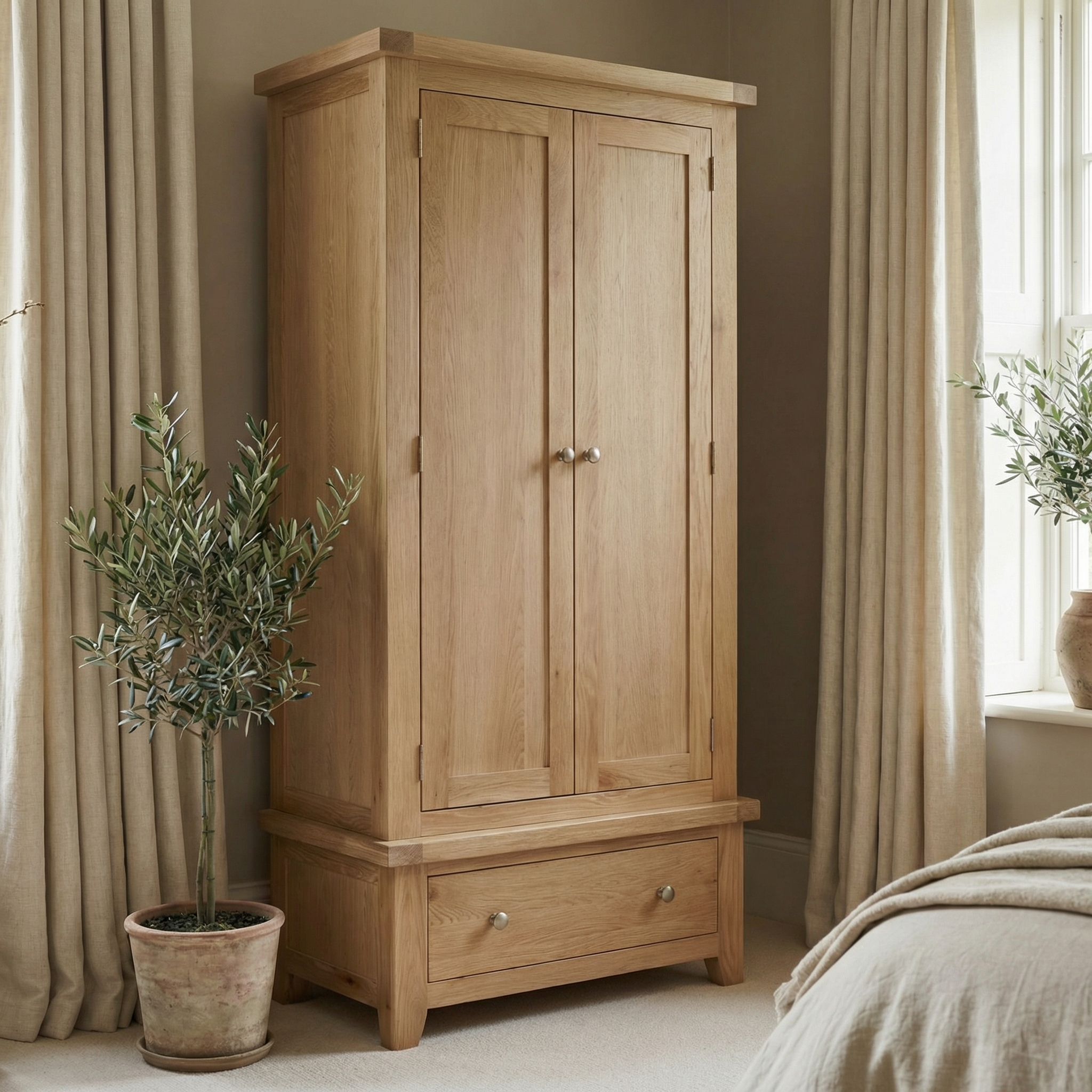 The Burford Double Wardrobe with Drawer in Natural Oak stands beside a potted plant in a neutral-toned bedroom, adding warmth and storage with its light wood finish and two-door design.