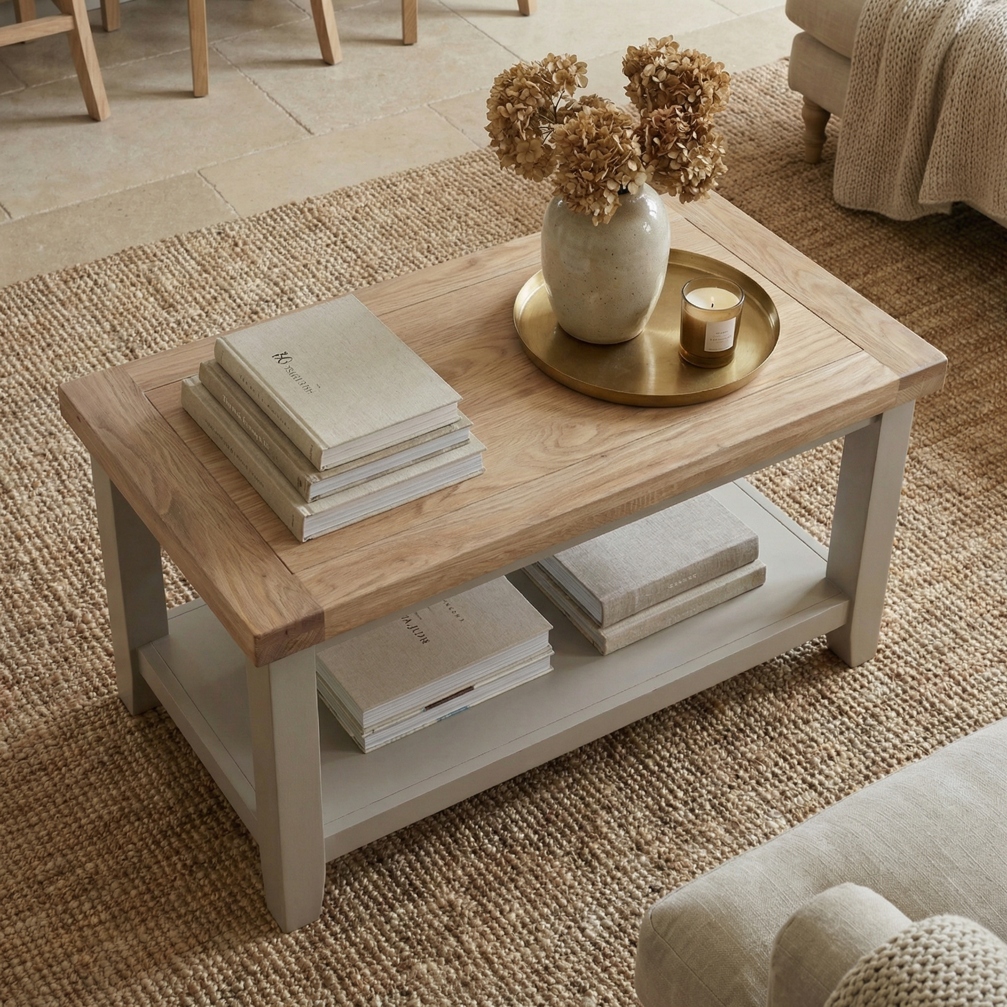 The Burford Coffee Table with Shelf in Pebble Grey offers space for stacking books, displaying a vase of dried flowers, and placing a candle on a gold tray.