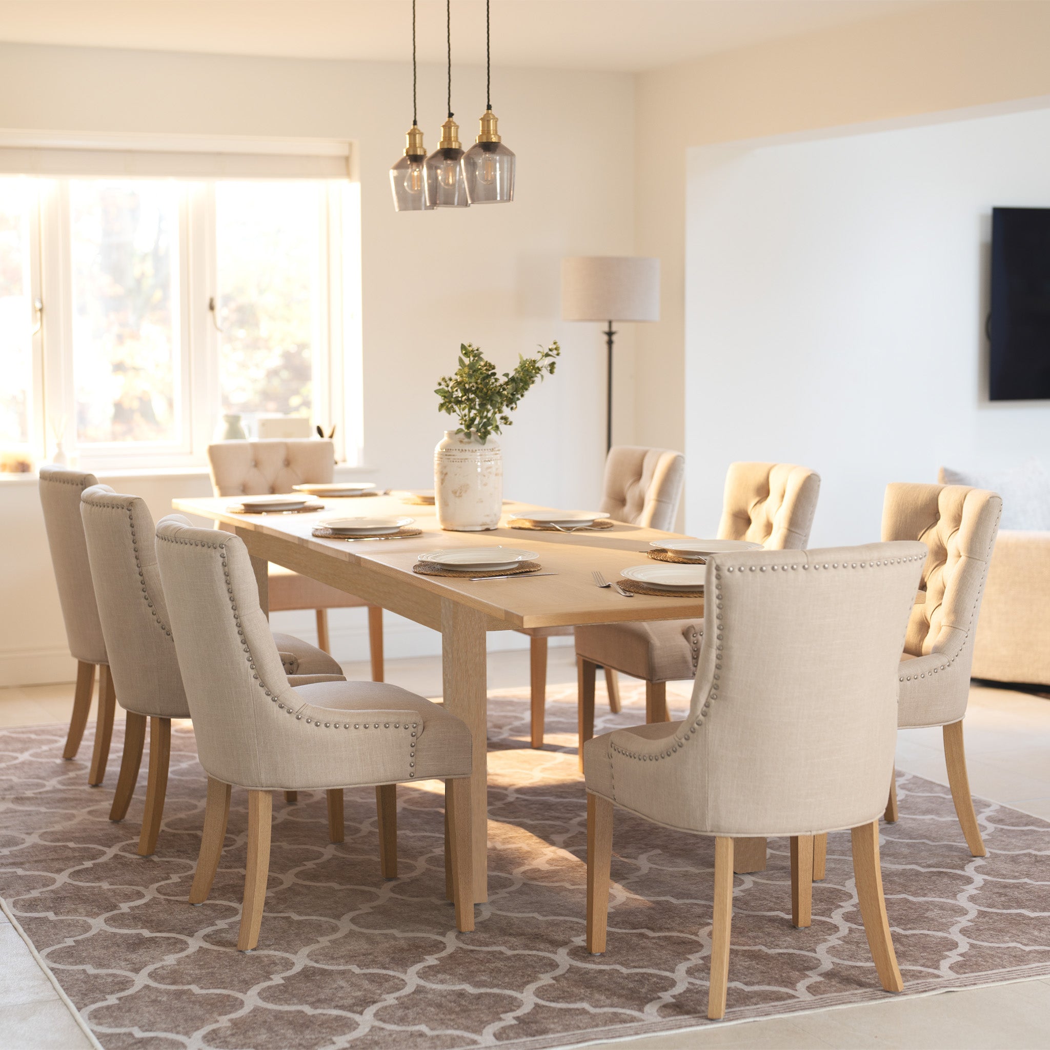 Bright dining room with a Burford 8 Seat Extending Dining Set, featuring a natural oak extendable table, eight Regent ivory chairs, and a patterned rug under natural light.