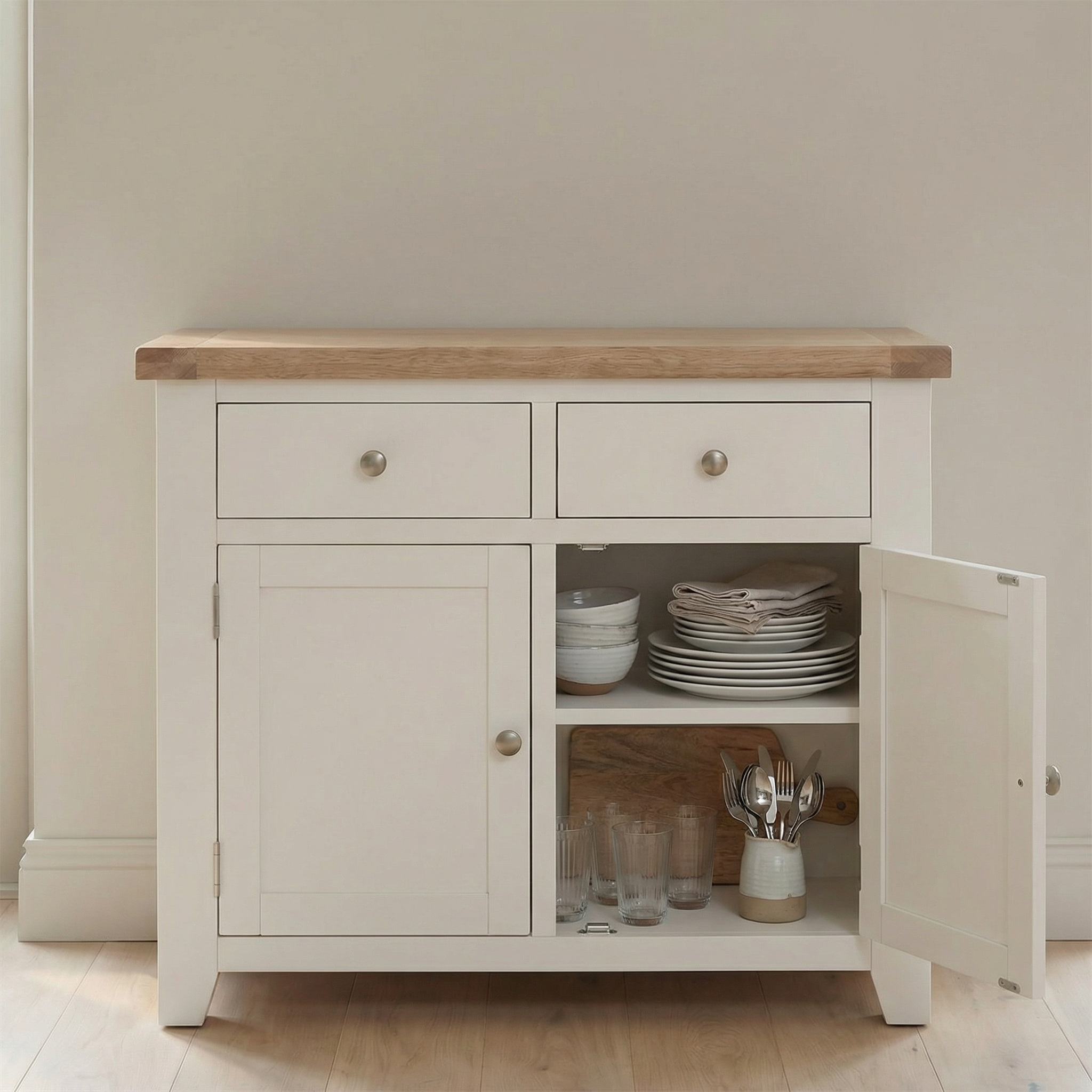 The Burford 2 Door 2 Drawers Sideboard in Warm White is shown with an open door, displaying neatly stacked plates, bowls, glasses, and utensils inside.
