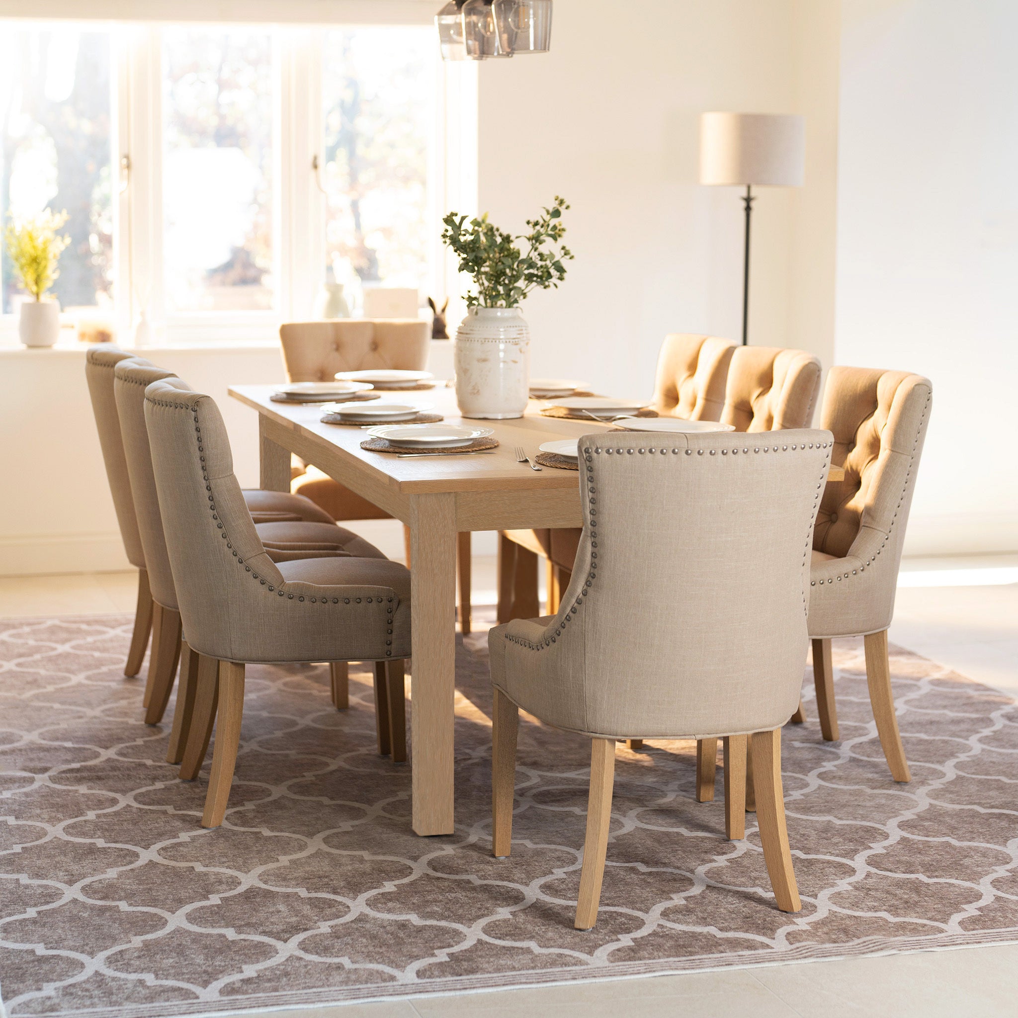 The Burford 8 Seat Extending Dining Set with Regent Chairs in Natural Oak and Beige, featuring eight upholstered chairs and an extendable table, brightens this sunlit dining room.