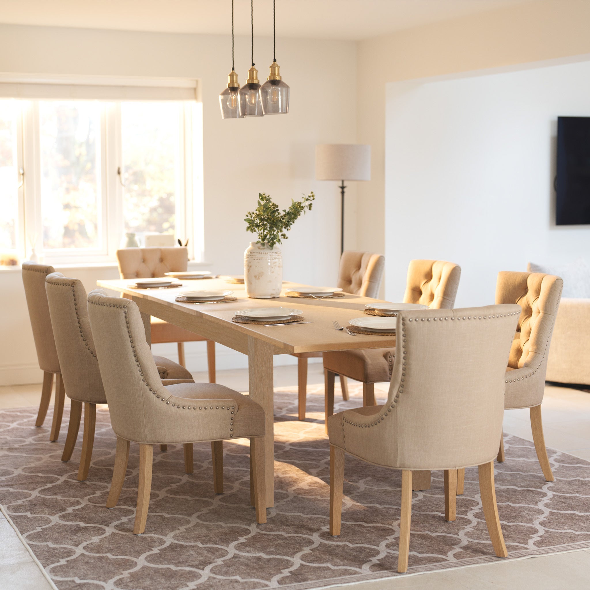 The Burford 8 Seat Extending Dining Set with Regent Chairs in natural oak and beige creates a warm, inviting atmosphere in the sunlit room, enhanced by tufted chairs and a patterned rug.