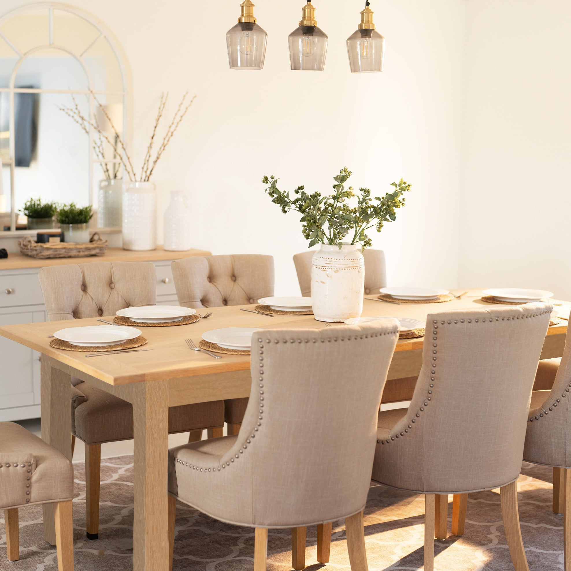 A bright dining room featuring the Burford 8 Seat Extending Dining Set with Regent Chairs in natural oak and beige, complete with a wooden extendable table and a decorative vase filled with greenery.