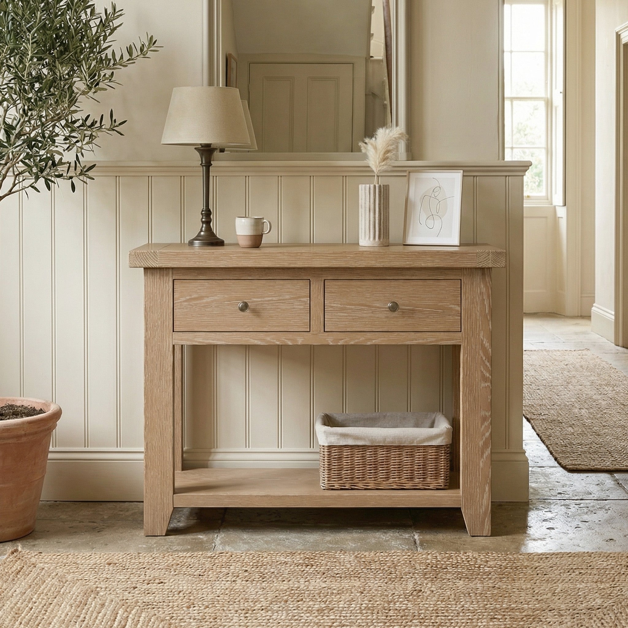 The Burford 2 Drawers Console Table in Natural Oak, styled with a lamp, decor, and a basket, elevates this bright, neutral hallway.