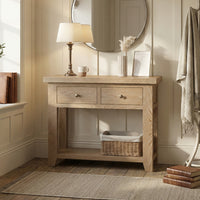 The Burford 2 Drawers Console Table in Natural Oak sets a warm tone in this bright room, styled with a lamp, cup, artwork, and a basket underneath—all complemented by cozy neutral decor.