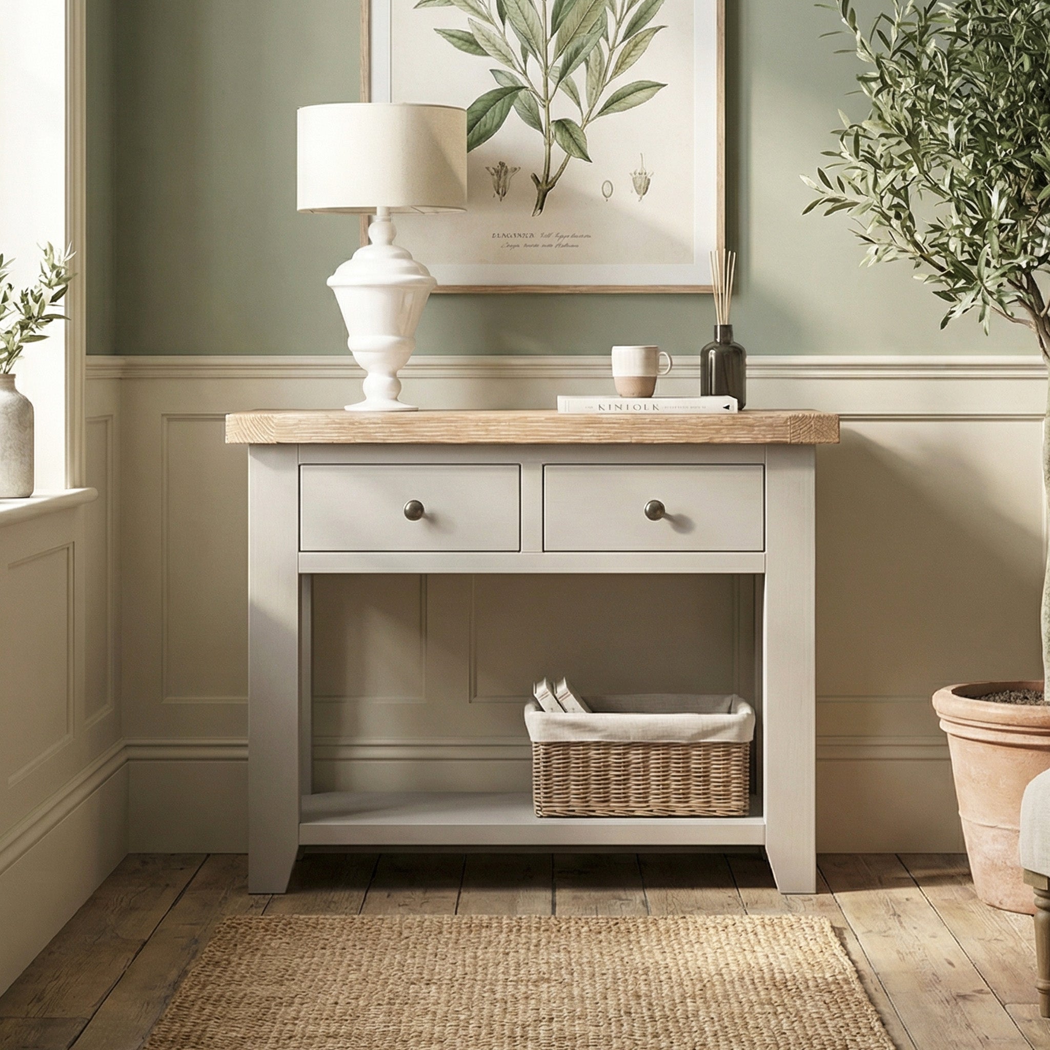 The Burford 2 Drawers Console Table in Pebble Grey sits under a botanical print, featuring two drawers and a basket on the shelf.
