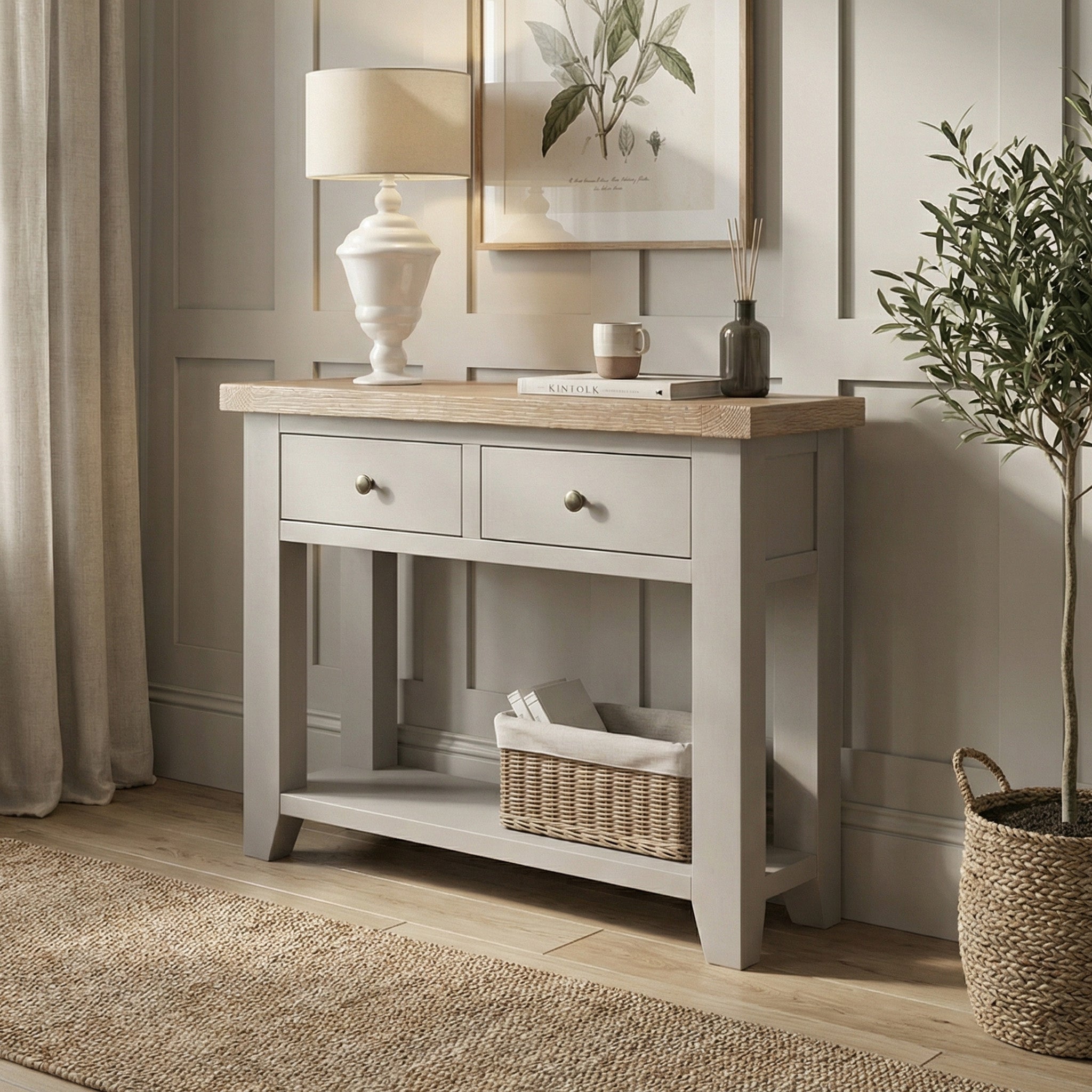 The Burford 2 Drawers Console Table in Pebble Grey, styled with decor, a lamp, a plant, and a basket on wooden flooring, enhances the cozy ambiance of this room.