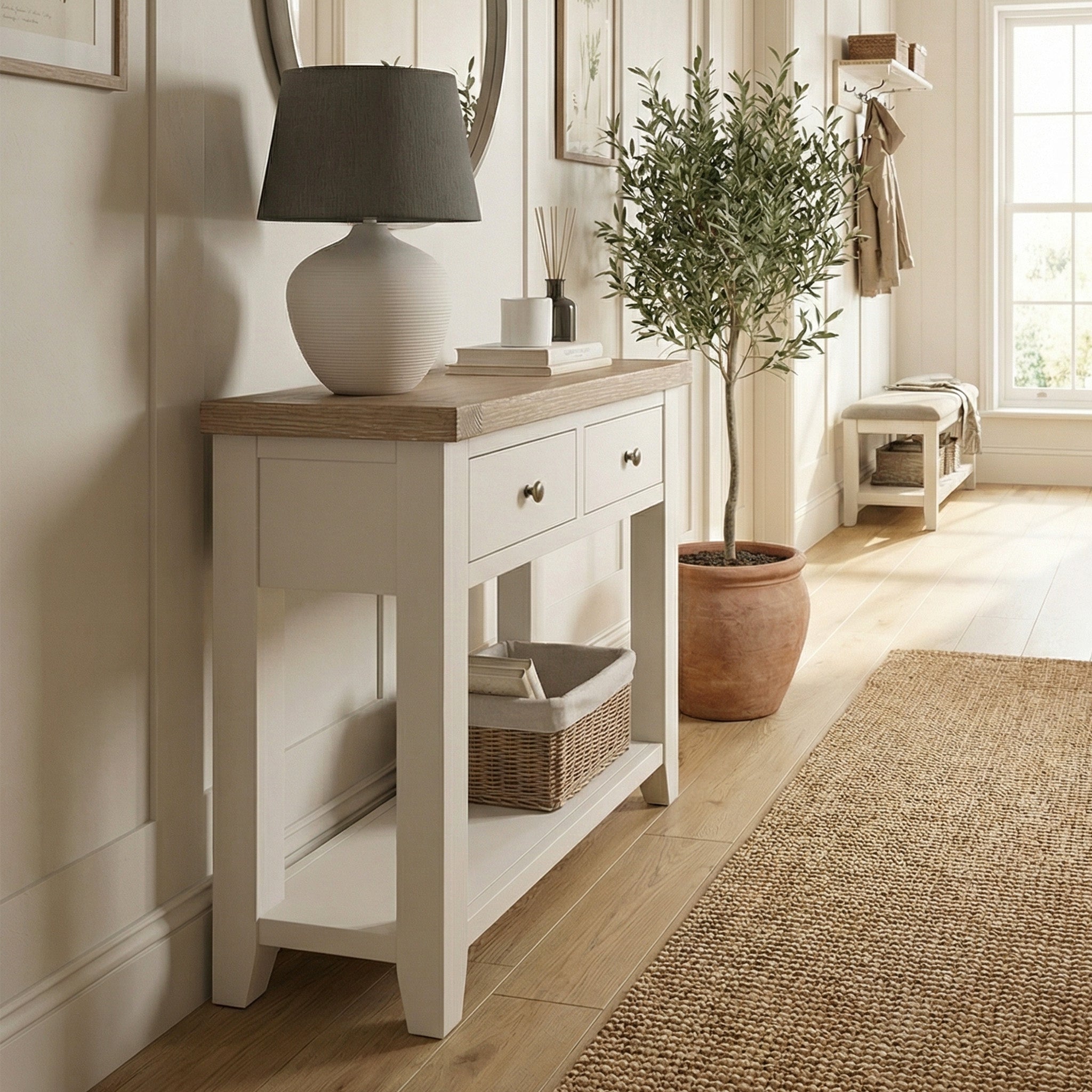 The Burford 2 Drawers Console Table in Warm White is styled with a gray lamp, basket, and potted plant, illuminated by sunlight streaming through a window.