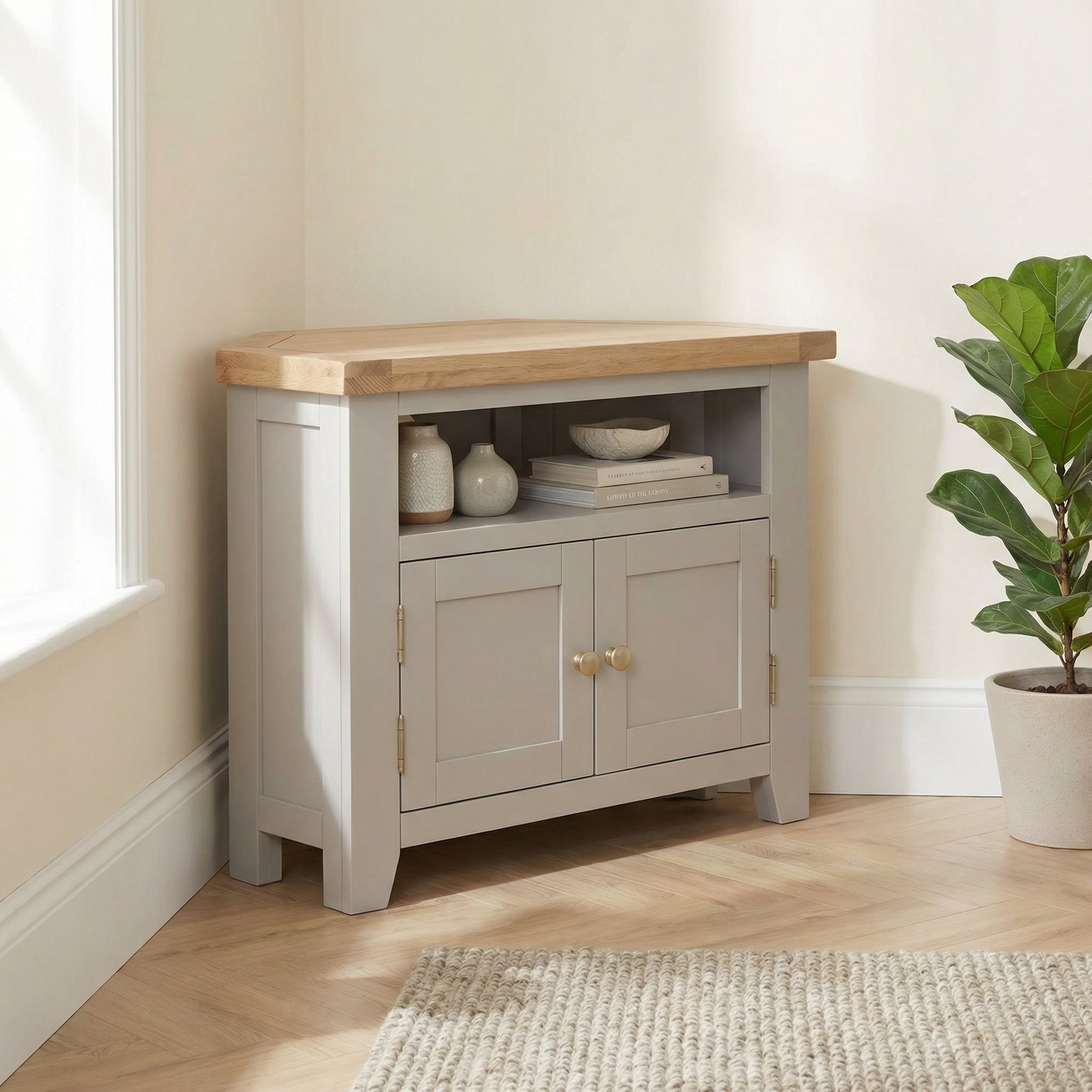 The Burford Corner Media Unit in Pebble Grey, featuring a natural oak top, books, and décor, sits beside a potted plant on a wooden floor, adding rustic charm to your space.
