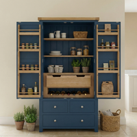 A Burford Double Larder Unit in Navy Blue with open doors displays shelves stocked with jars, woven baskets, wine bottles, and kitchen storage essentials, set amid greenery.