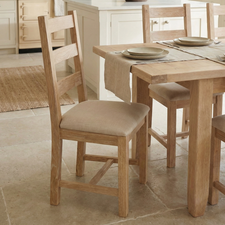 Light wood dining table with Burford Set of 2 Ladderback Dining Chairs with Fabric Seat in Natural Oak, featuring beige cushions and set with plates in a bright kitchen.