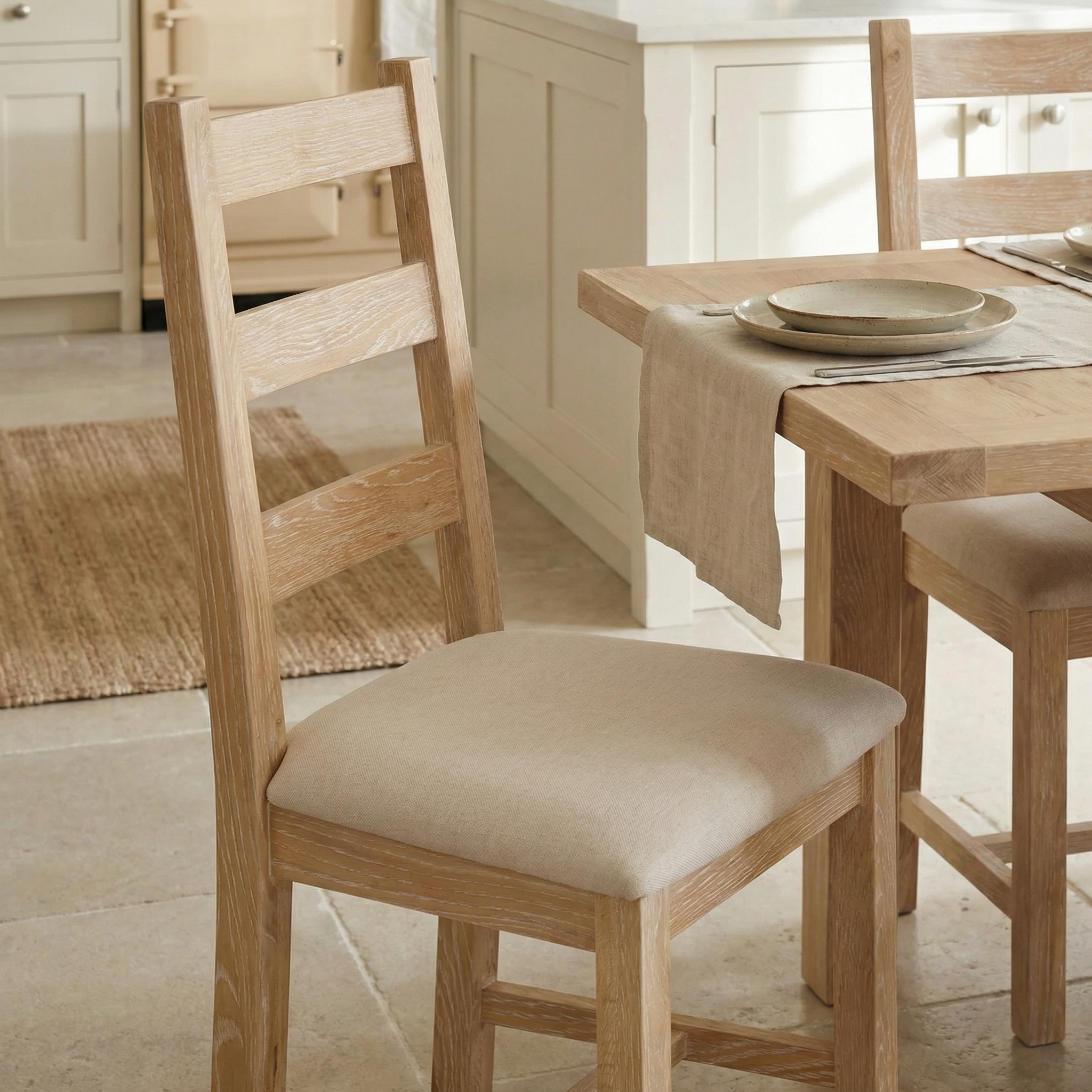 The Burford Set of 2 Ladderback Dining Chairs with Fabric Seat in Natural Oak surrounds a matching table in a bright kitchen.