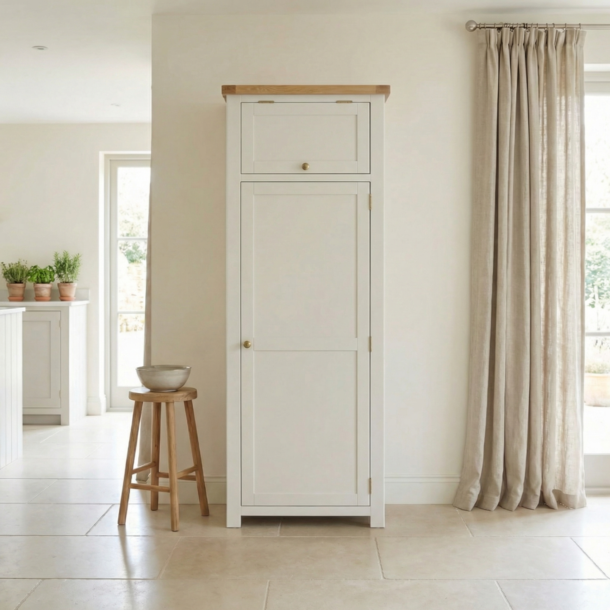 The Burford Single Larder Unit in Warm White with a Natural Oak top stands beside a wooden stool and beige curtains in a bright, airy room.