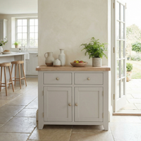 The Burford 2 Door 2 Drawers Sideboard in Pebble Grey, styled with decor and a plant, stands in a bright kitchen featuring open doors leading to a garden.
