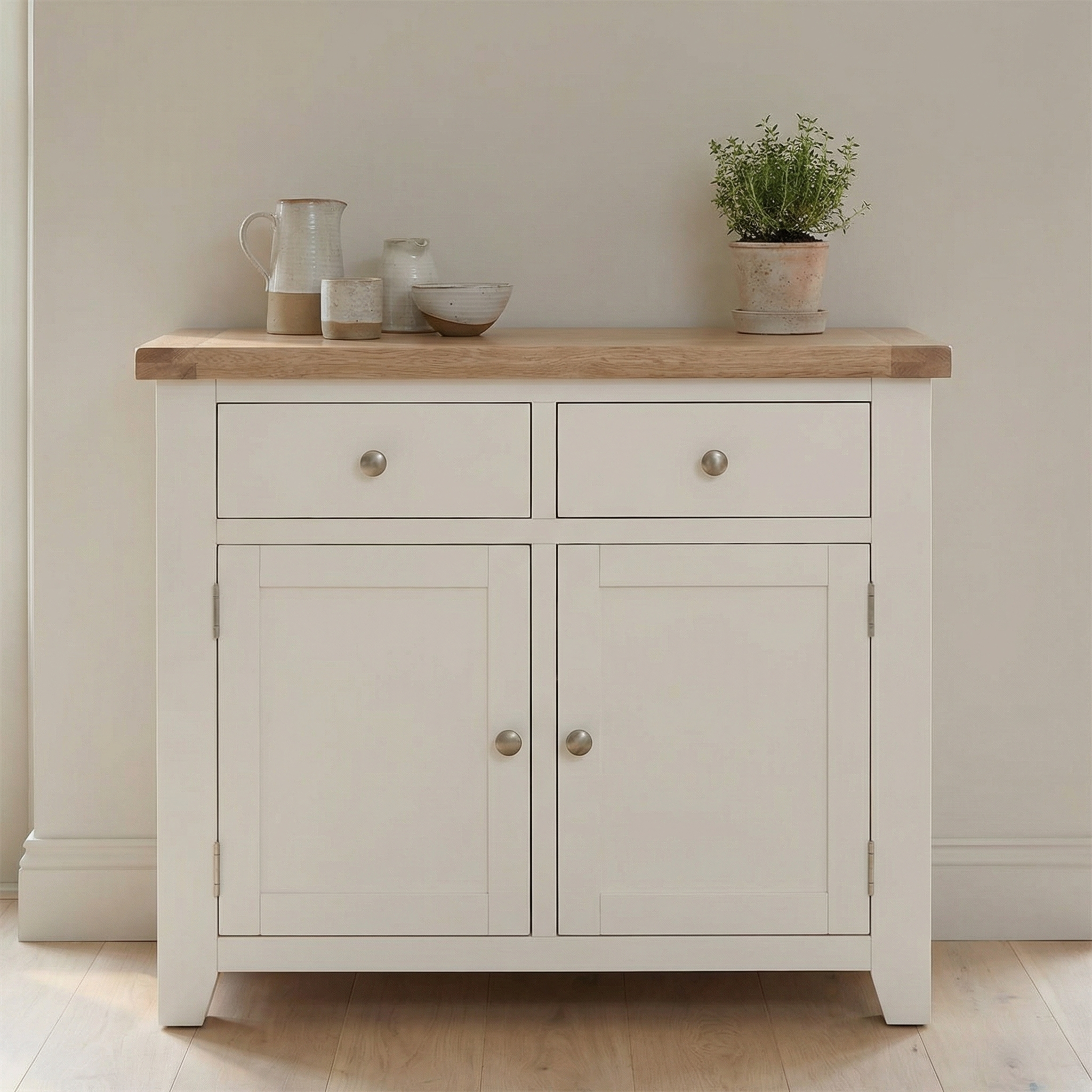 The Burford 2 Door 2 Drawers Sideboard in Warm White features a wooden top, spacious drawers, and cabinets. Styled with décor like a plant, bowls, and a pitcher, it creates a welcoming look for any room.