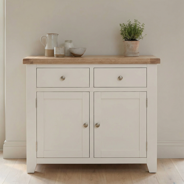 The Burford 2 Door 2 Drawers Sideboard in Warm White features a wooden top, spacious drawers, and cabinets. Styled with décor like a plant, bowls, and a pitcher, it creates a welcoming look for any room.