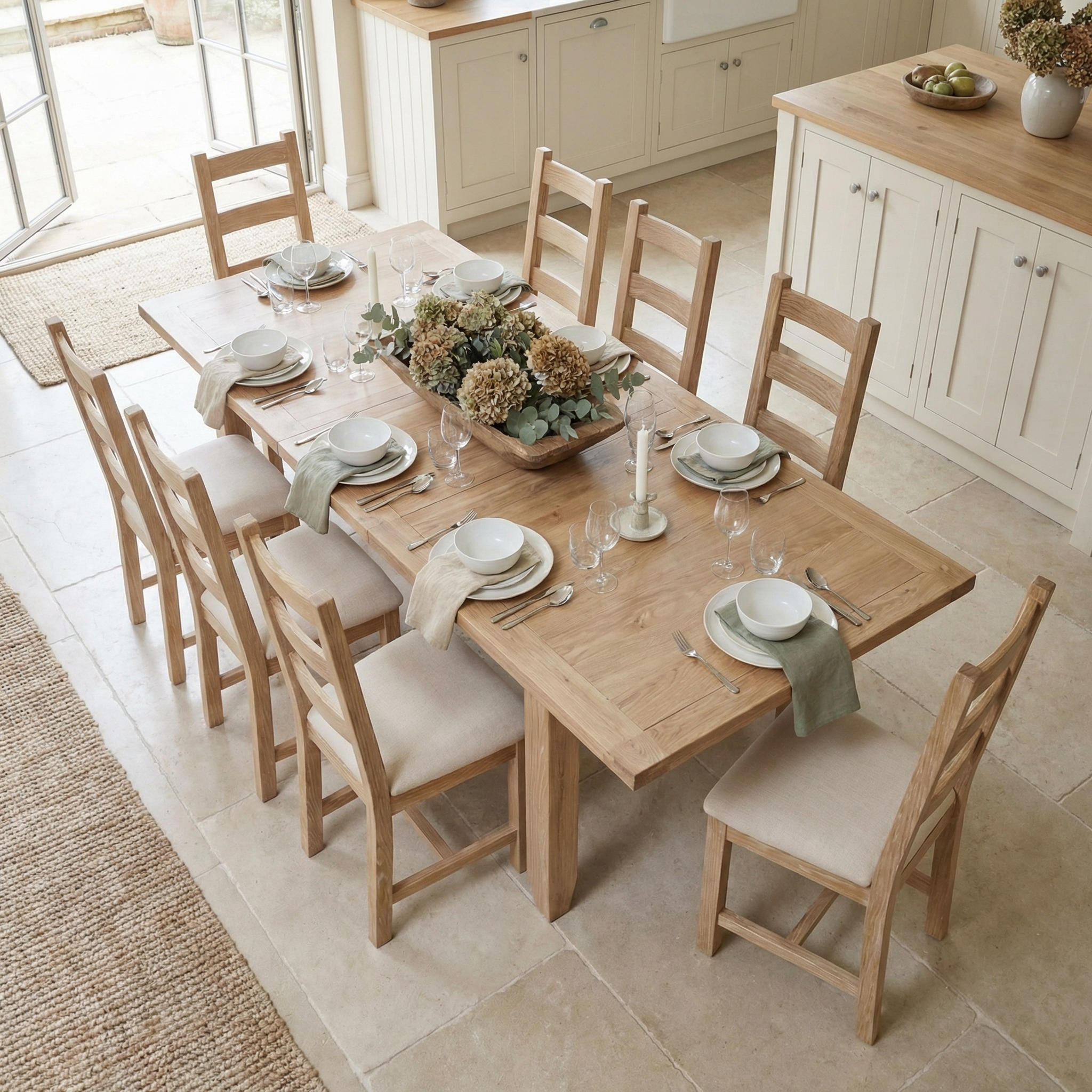 The Burford 8 Seat Extending Dining Set in Natural Oak adds elegance to a bright kitchen, complete with plates, glasses, and a floral centerpiece—perfect for gatherings.