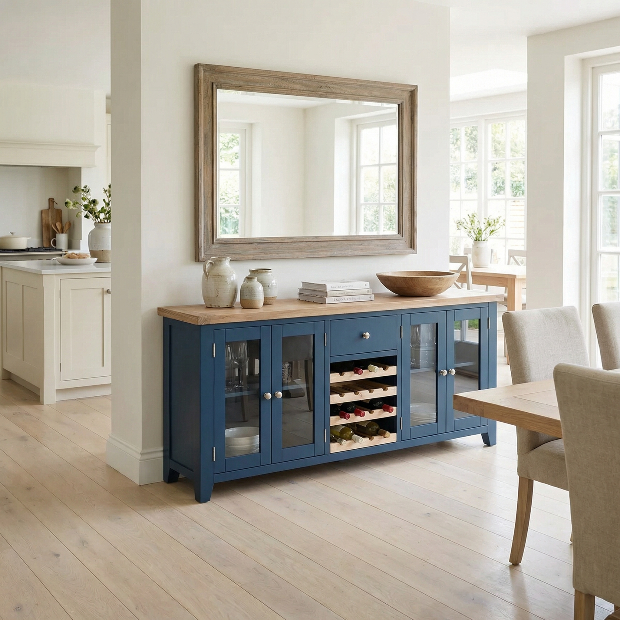 The Burford Grand Sideboard with Wine Rack in Navy Blue is placed against a wall with a large mirror in a bright dining room. Its glass doors and built-in wine rack add both style and storage for decor.