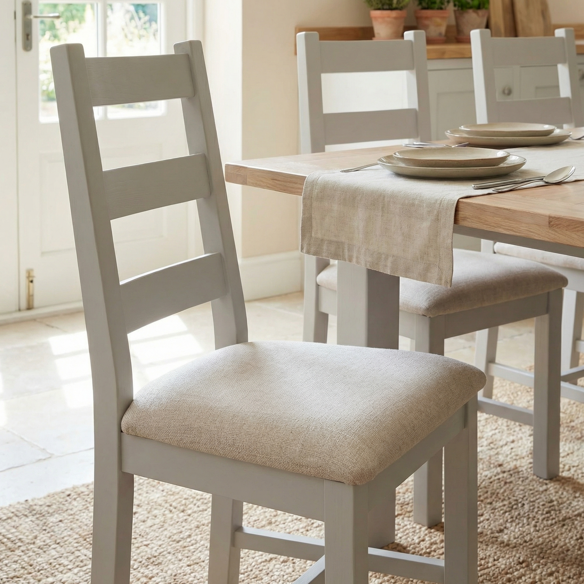 A Burford Set of 2 Ladderback Dining Chairs with Fabric Seat in Pebble Grey is placed at a dining table set for a meal in a bright, airy room.