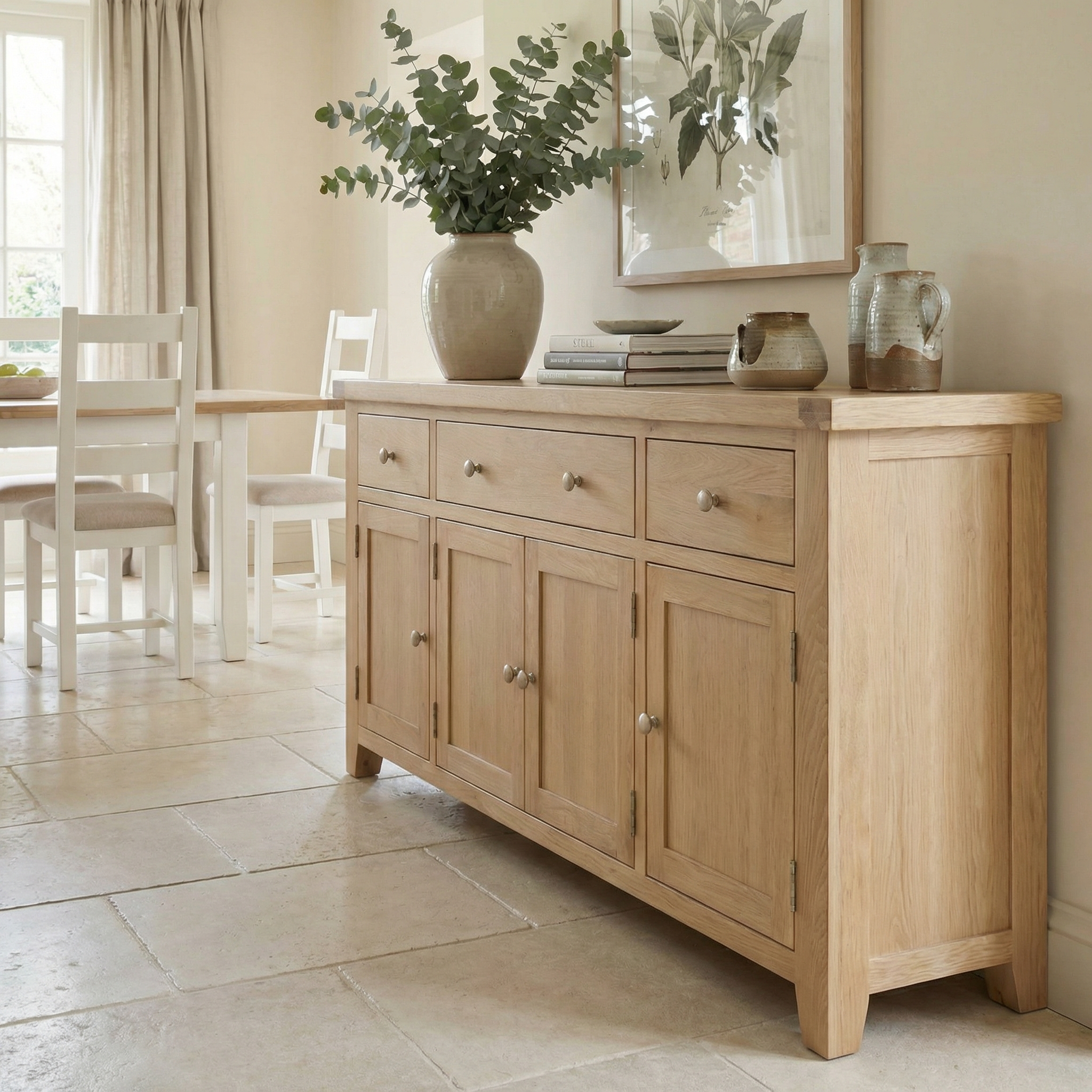 The Burford Large 4 Door 3 Drawers Sideboard in Natural Oak is styled with a vase, books, and pottery in a bright dining room with neutral decor and a tiled floor.