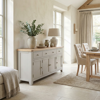 Light-filled dining room featuring the Burford Large 4 Door 3 Drawers Sideboard in Pebble Grey, neutral decor, a large vase with greenery, and upholstered chairs.