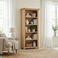 The Burford Large Bookcase in Natural Oak stands with decor items in a bright, cozy living room featuring white curtains and a beige armchair.