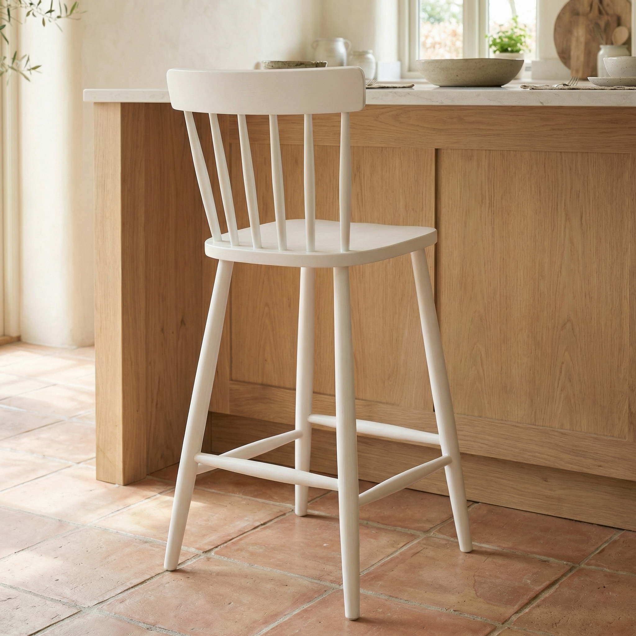 A Burford Spindleback Bar Stool in Warm White stands by a light wood kitchen island, providing stylish seating on a tiled floor.