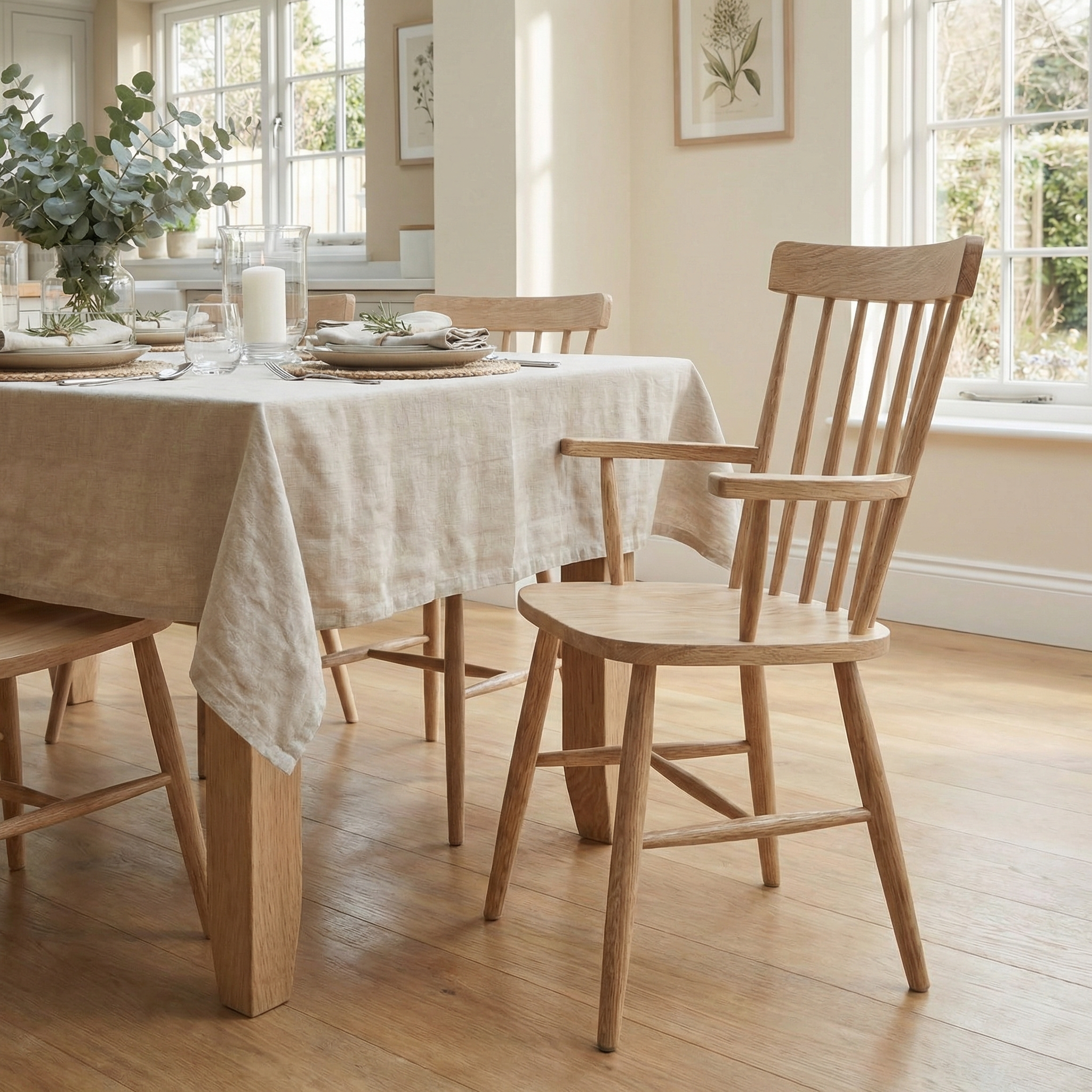A sunlit room features a wooden dining table with a beige tablecloth, set for a meal, and Burford Spindleback Dining Chairs with Armrest in Natural Oak.