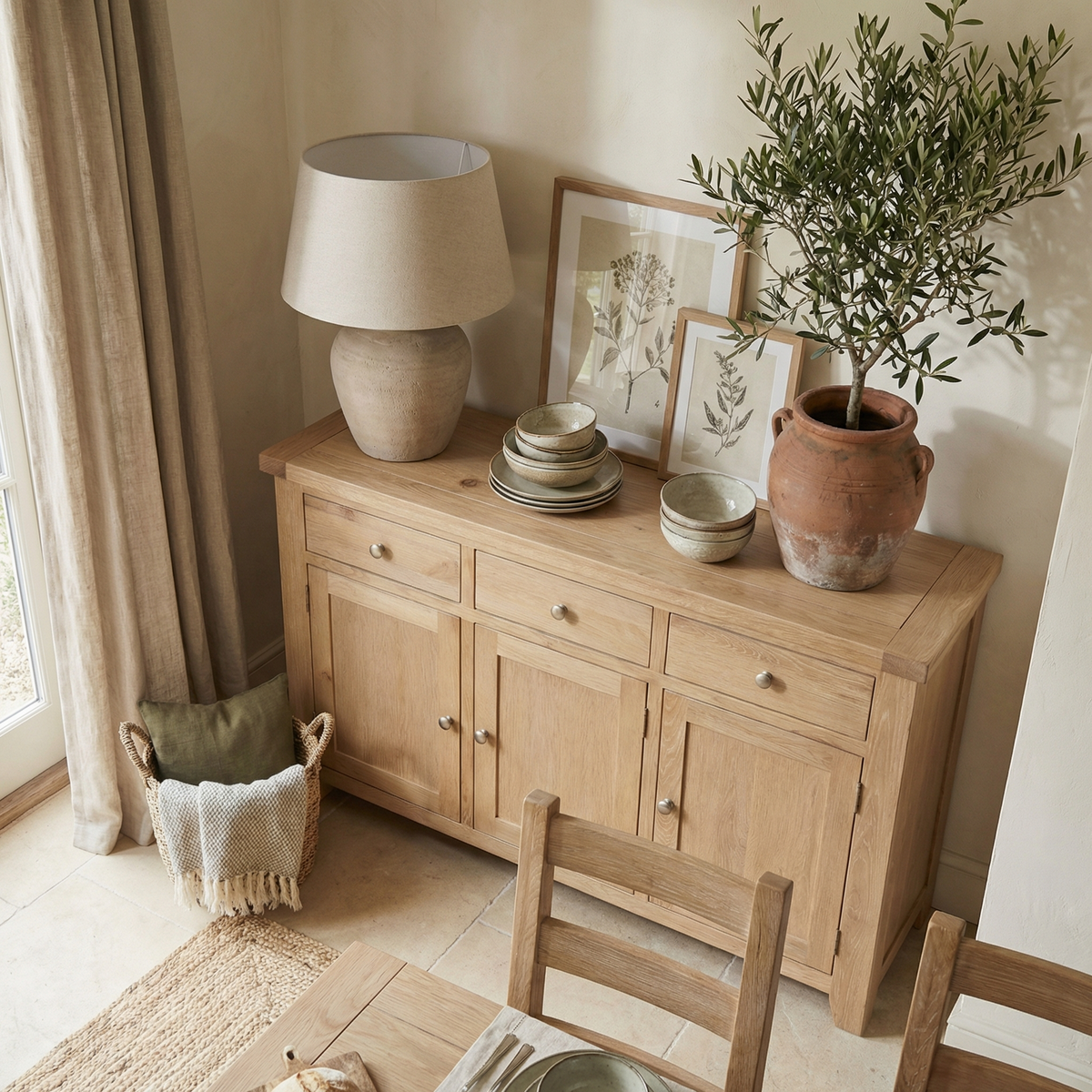 The Burford Large 3 Door 3 Drawers Sideboard in Natural Oak, styled with a lamp, potted plant, framed art, stacked plates, and bowls, creates a cozy dining room ambiance.