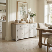 Light-filled dining room featuring the Burford Large 3 Door 3 Drawers Sideboard in Pebble Grey, vases, framed botanical art, and a wooden table with chairs.