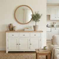 The Burford Large 3 Door 3 Drawers Sideboard in Warm White, styled with pottery, a potted plant, and a round mirror, adds warmth and elegance to this neutral, minimalist living space with its oak top and modern design.
