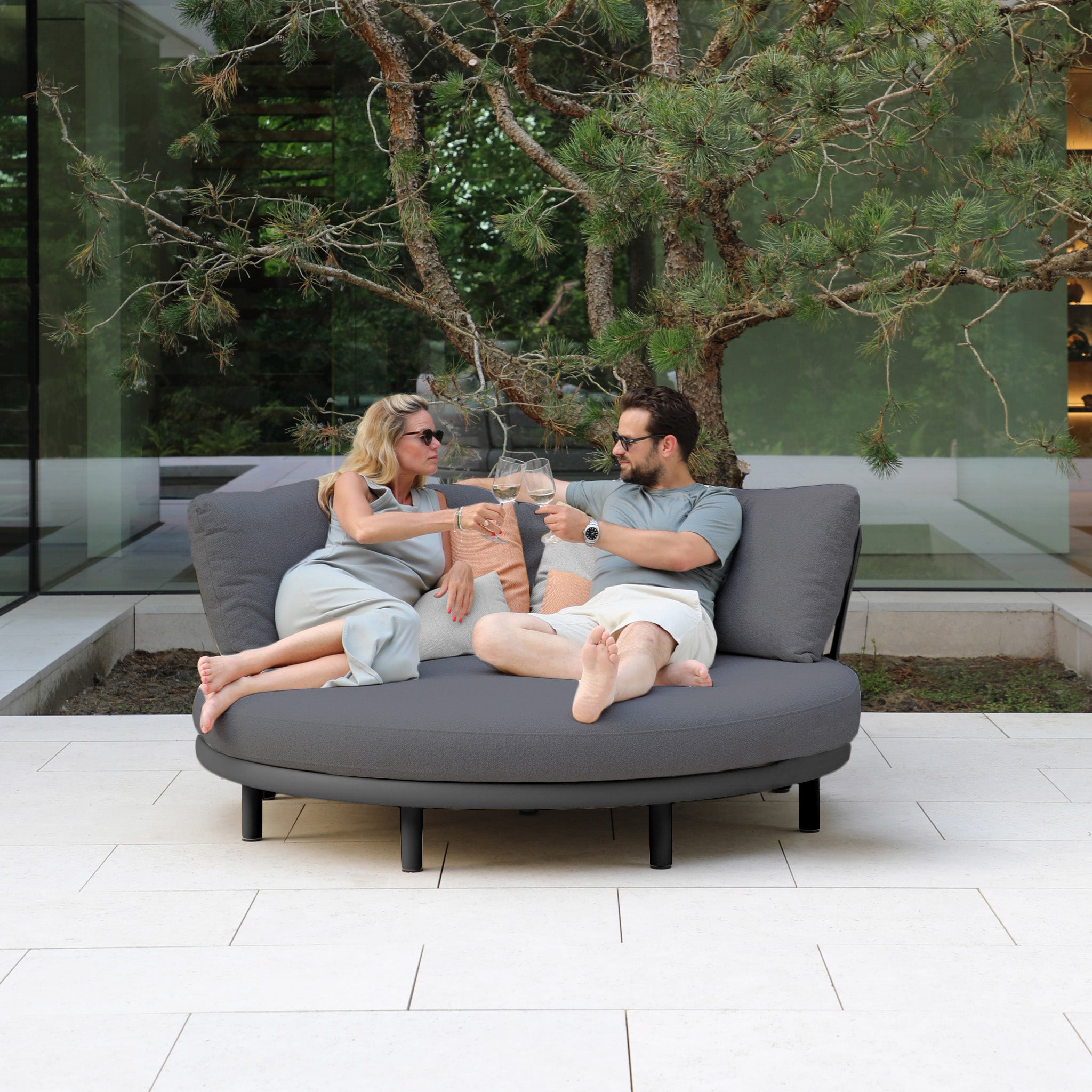 A couple relaxes and toasts glasses on a weather-resistant Selene Daybed in Charcoal, nestled amid trees and glass walls, embodying outdoor luxury.