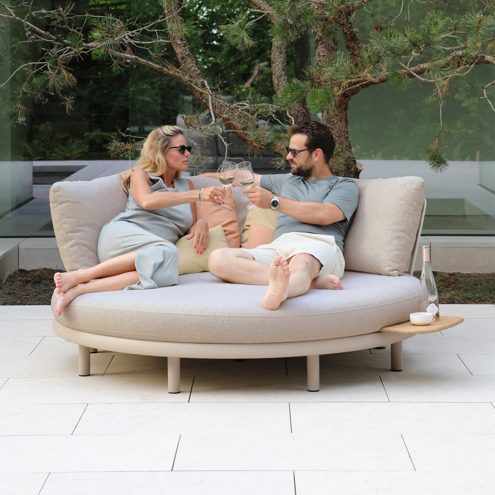 Two people relaxing on the weather-resistant Selene Daybed in Fawn, toasting with glasses and savoring outdoor luxury amidst lush greenery.