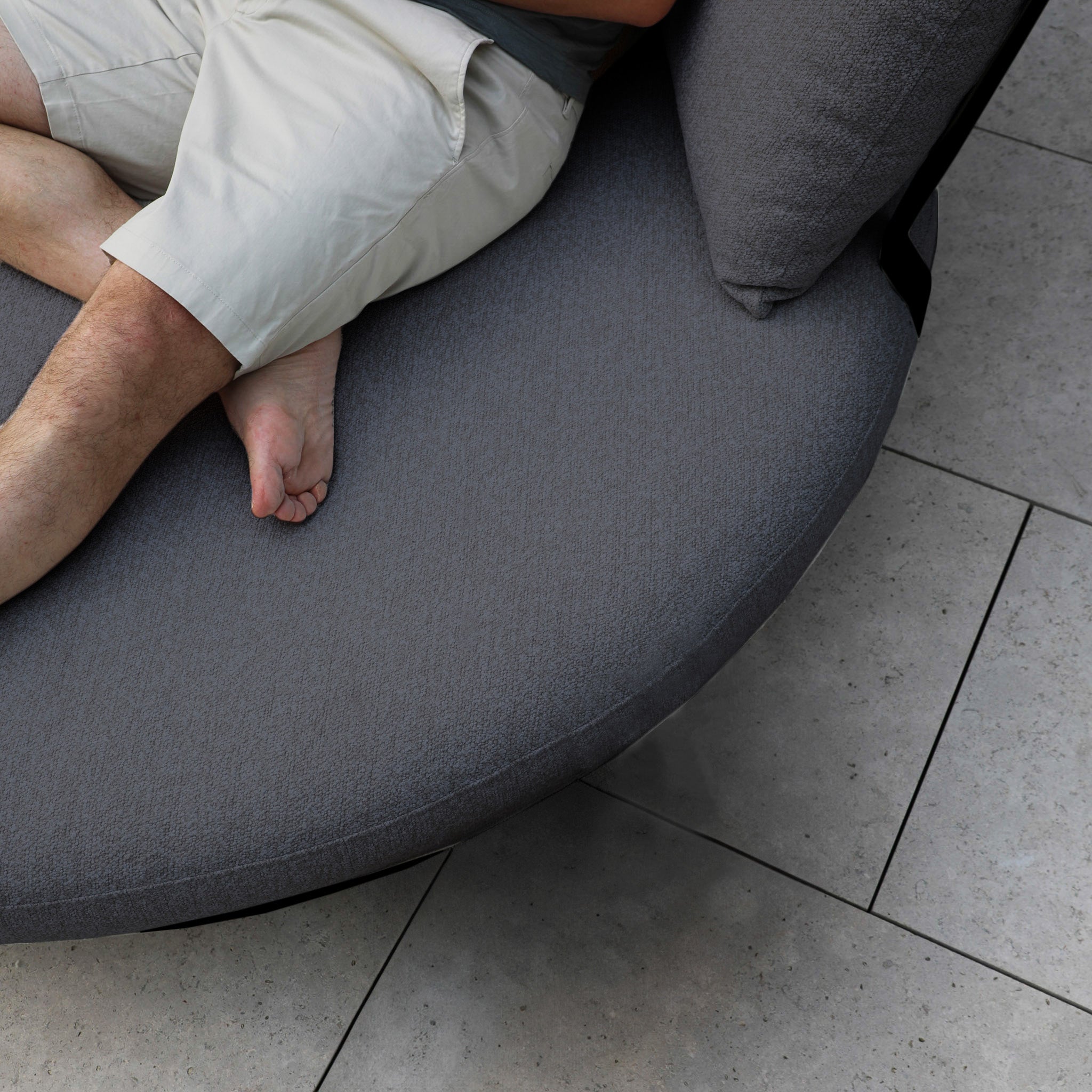 A person relaxes on a Selene Daybed in Charcoal, placed on a smooth tiled floor. They're dressed in light pants with one bare foot visible, showcasing a seamless mix of comfort and style in an elegant outdoor setting.