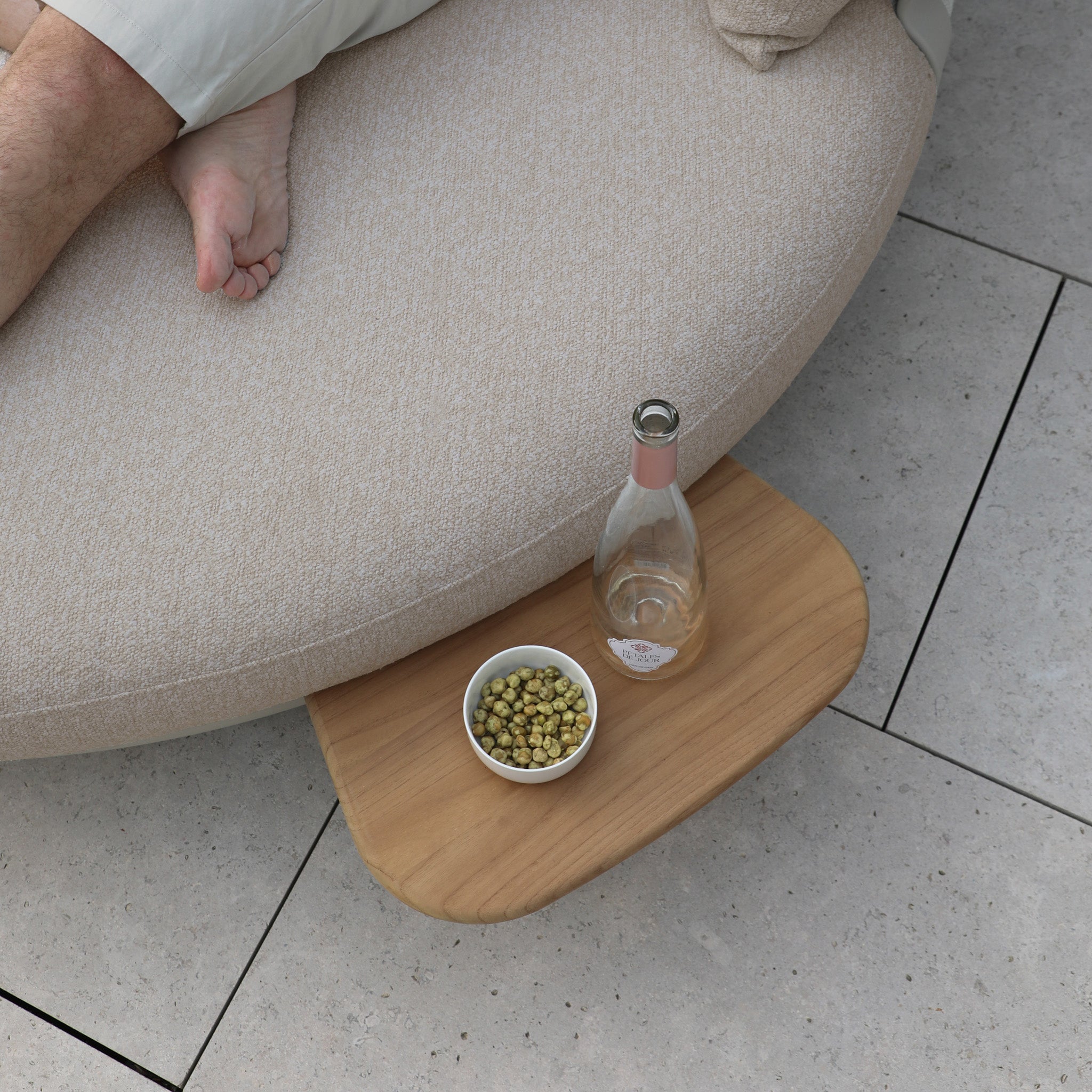 A person relaxes on the Selene Daybed in Fawn, a true symbol of outdoor luxury, with a table beside them holding a bottle and a bowl of snacks.