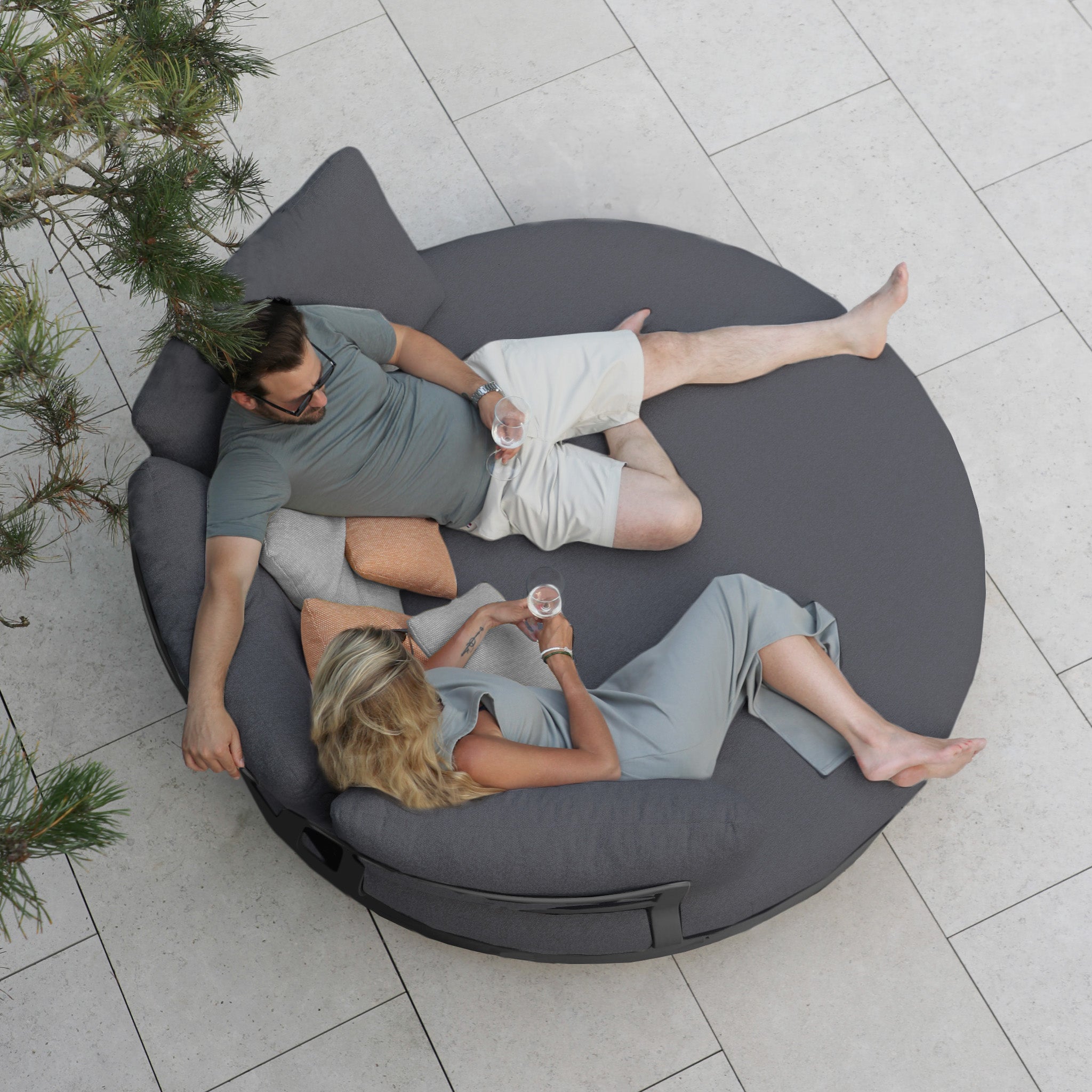 A man and woman relax on a Selene Daybed in Charcoal, sipping drinks and chatting under a tree on a tiled patio, embodying outdoor luxury with its weather-resistant design.