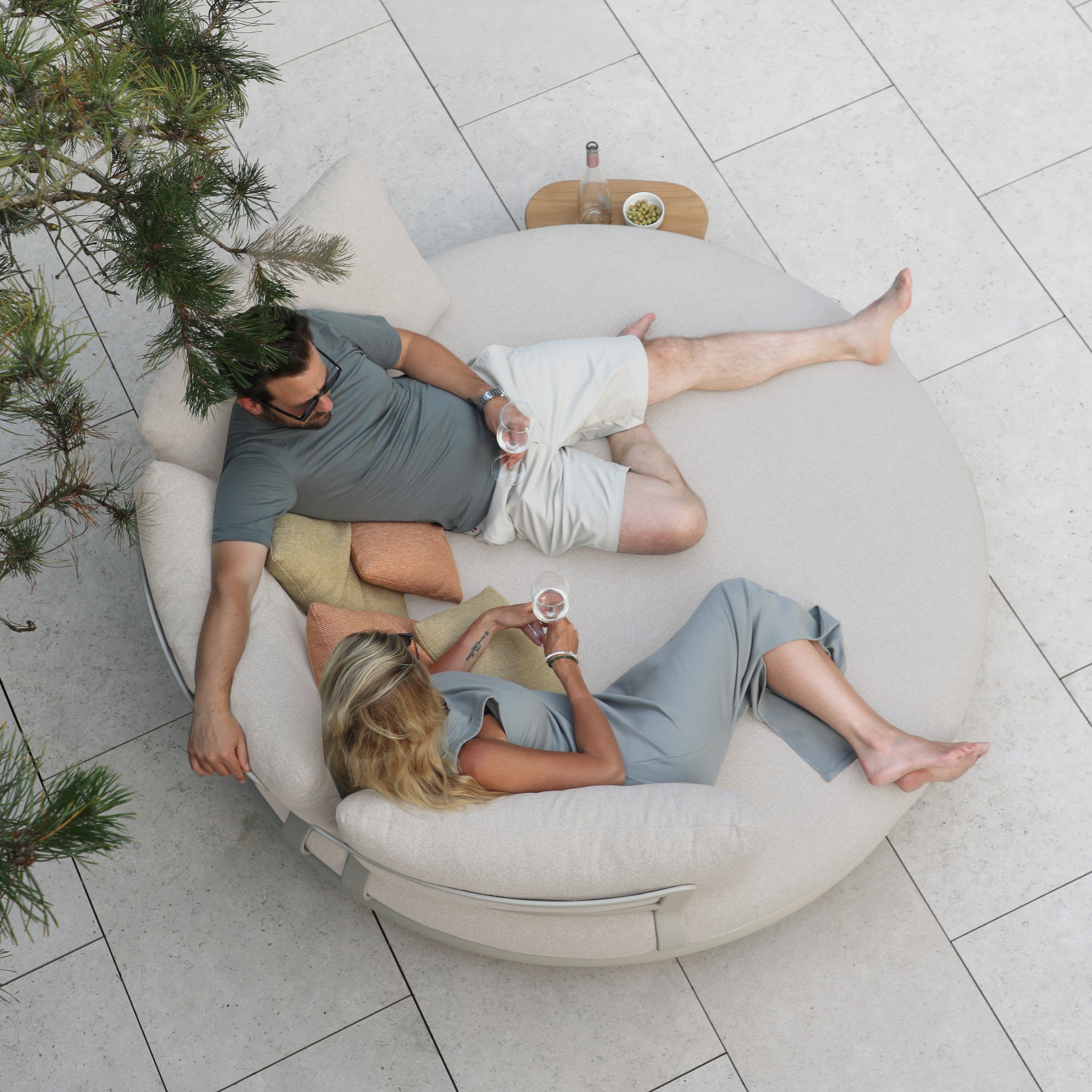 Two people relax with drinks on a Selene Daybed in Fawn under a tree, seen from above on a tiled patio. The weather-resistant design enhances their outdoor luxury experience.