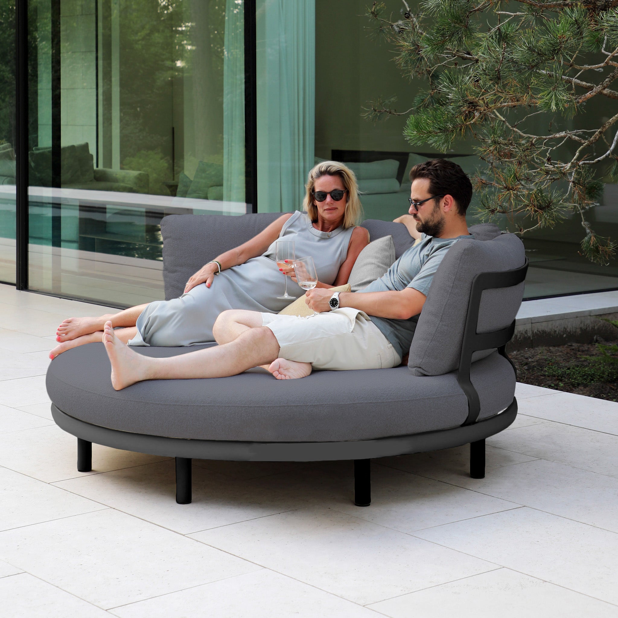 Two people relax on a Selene Daybed in Charcoal by a glass-walled building, enjoying their drinks and the stunning view. The luxurious outdoor setting combines comfort and style, with weather-resistant features for lasting relaxation.