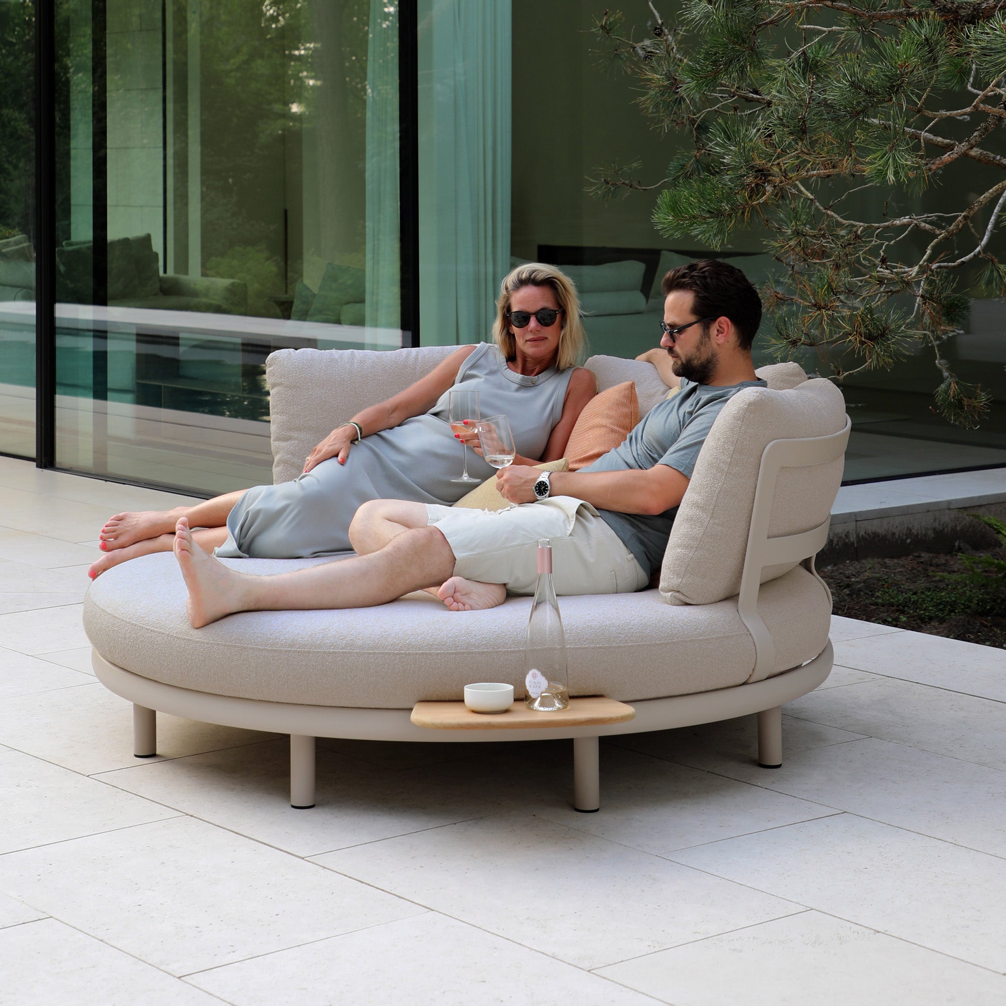 A couple relaxes on the sleek Selene Daybed in Fawn, a luxurious outdoor staple, sipping wine by the pool. Framed by glass windows, they revel in their weather-resistant sanctuary.