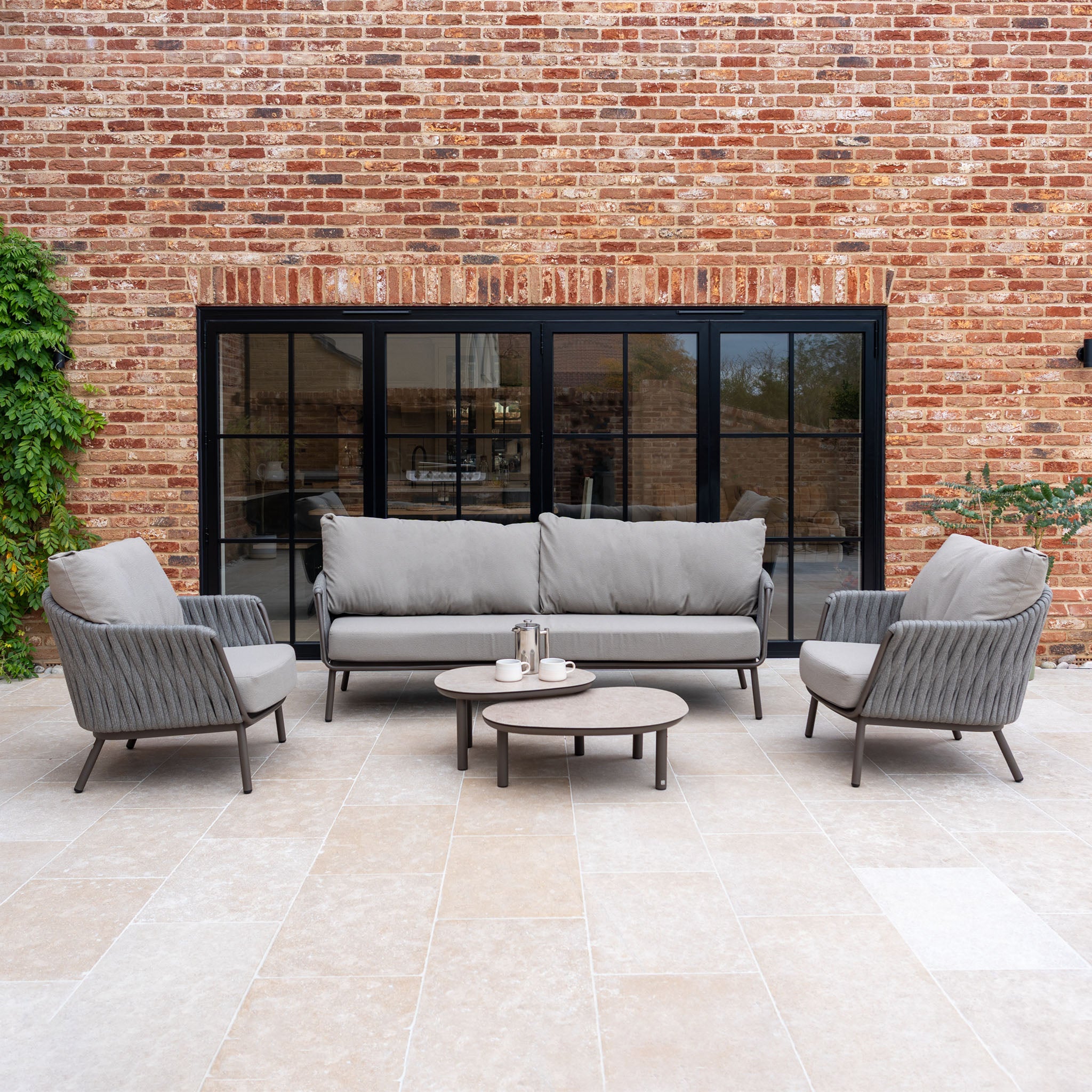 Modern outdoor patio featuring the Capri 3 Seat Sofa Set in Stone Grey with a round coffee table and chic brick wall backdrop—ideal garden furniture for relaxing or entertaining outdoors.