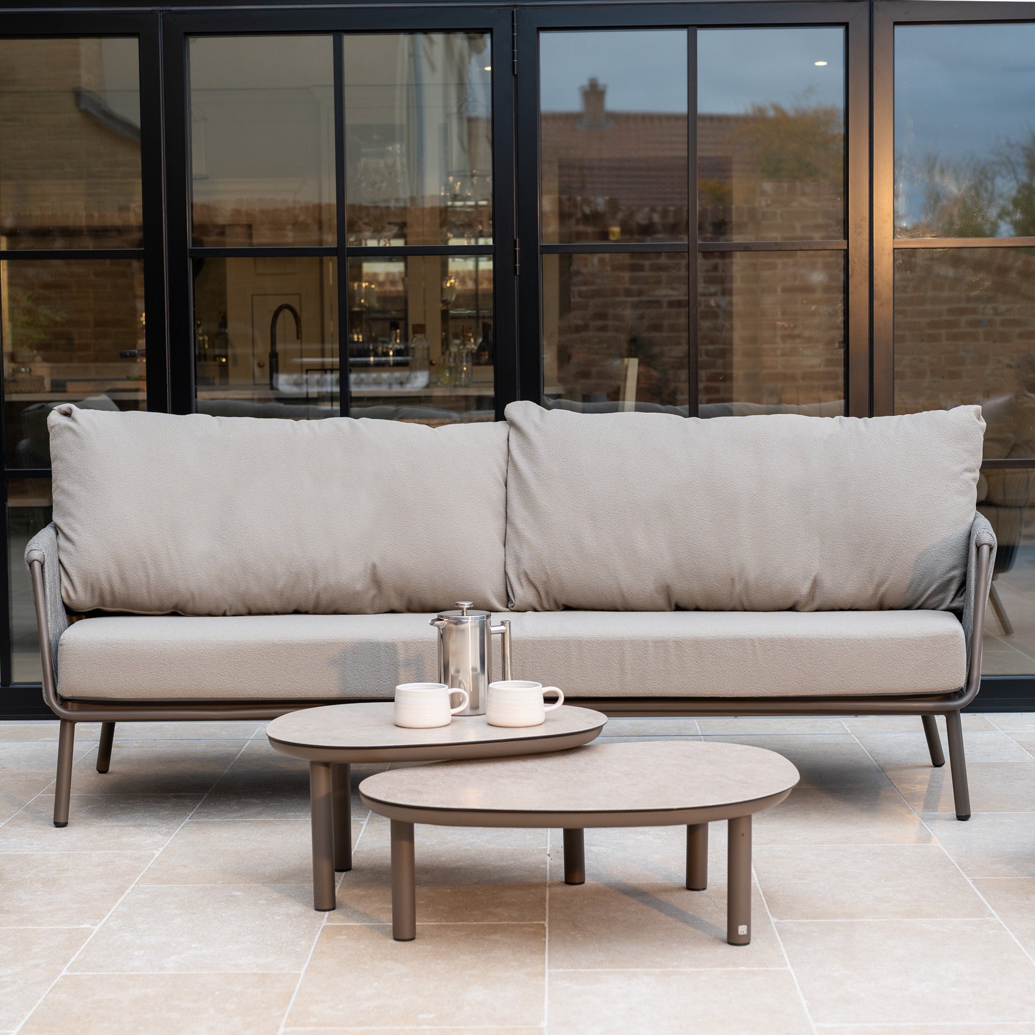 Capri 3 Seat Sofa Set in Stone Grey with two round coffee tables, coffee pot, and cups on a tiled patio in front of glass doors—perfect garden furniture for stylish outdoor living.