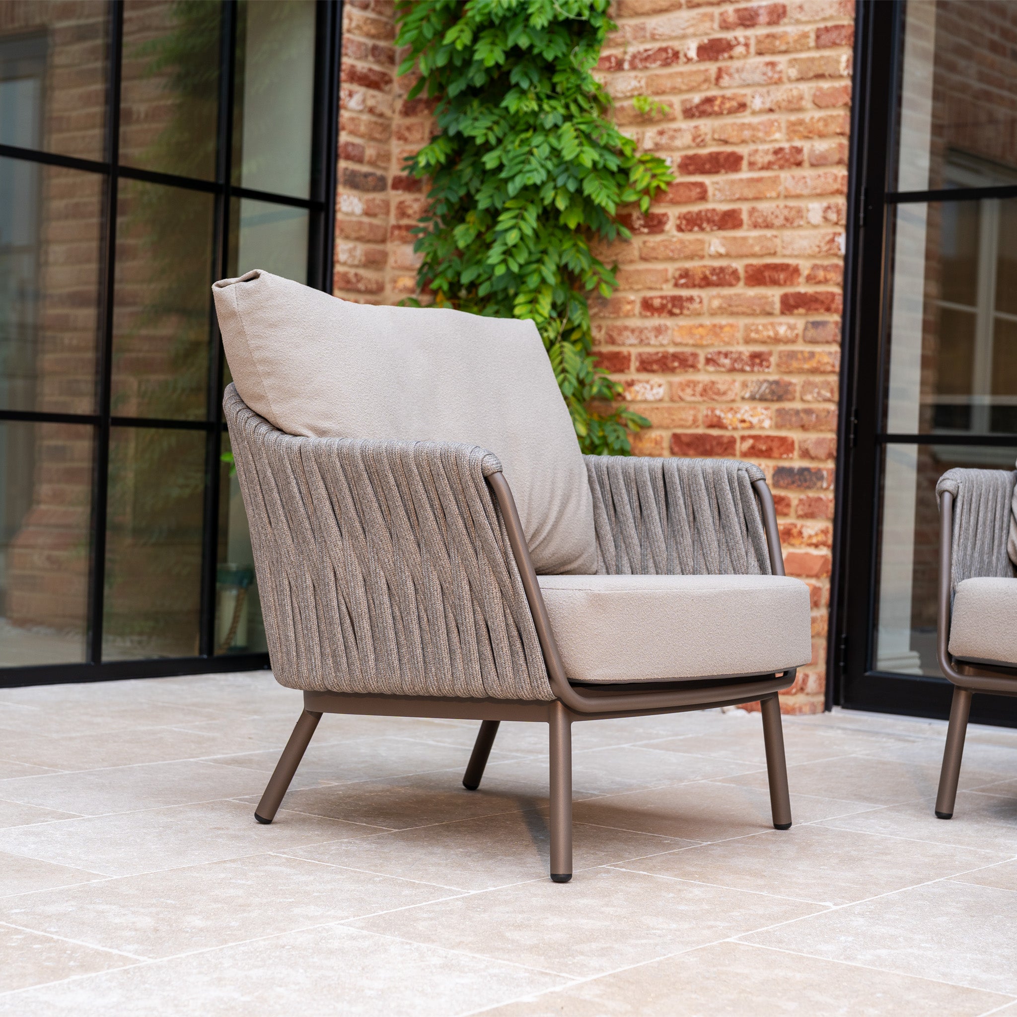 The Capri 3 Seat Sofa Set in Stone Grey adds elegance to a tiled patio with brick walls and green vines in the background.