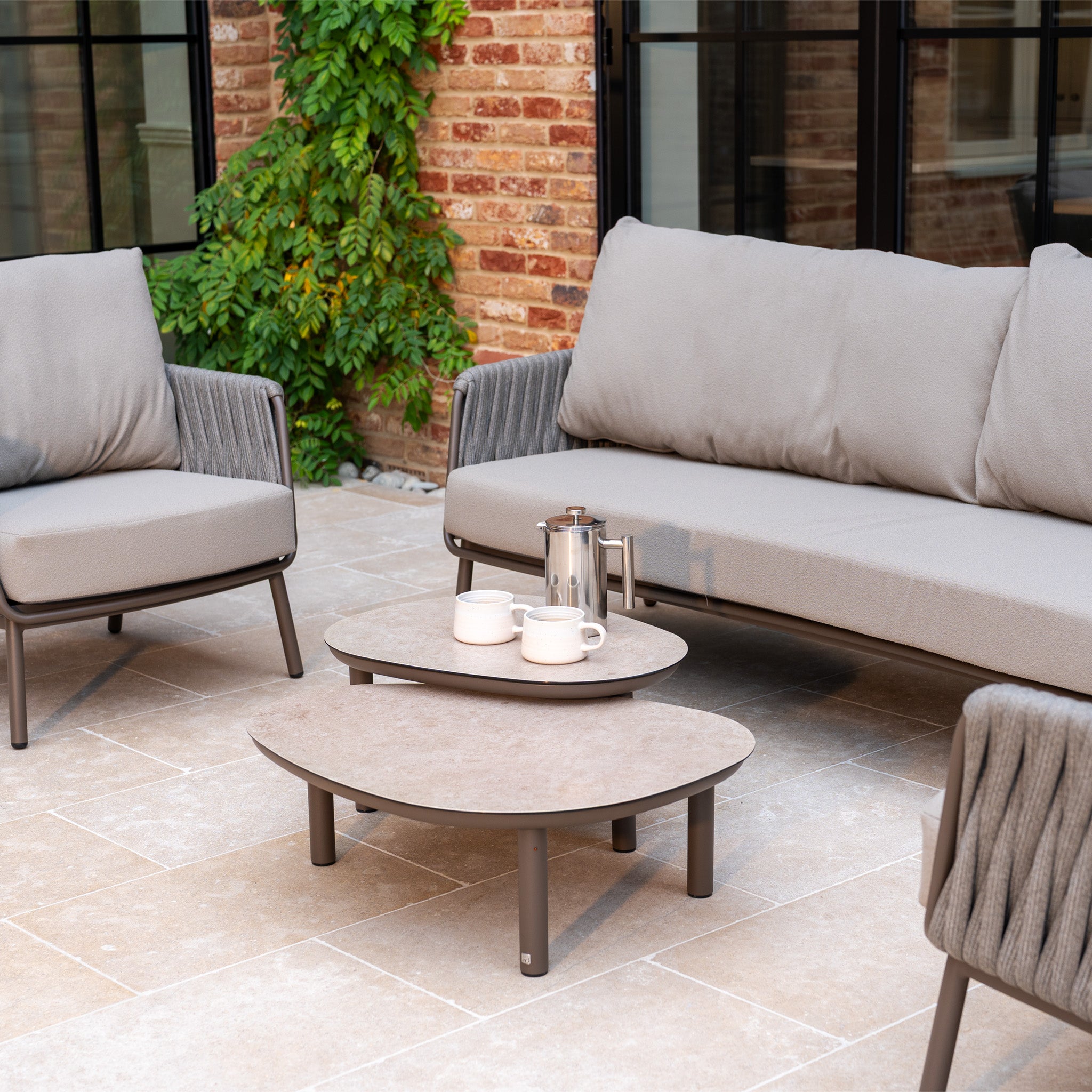 Modern outdoor patio featuring the Capri 3 Seat Sofa Set in Stone Grey and stylish garden furniture, complete with a coffee table and coffee set on beige stone flooring.
