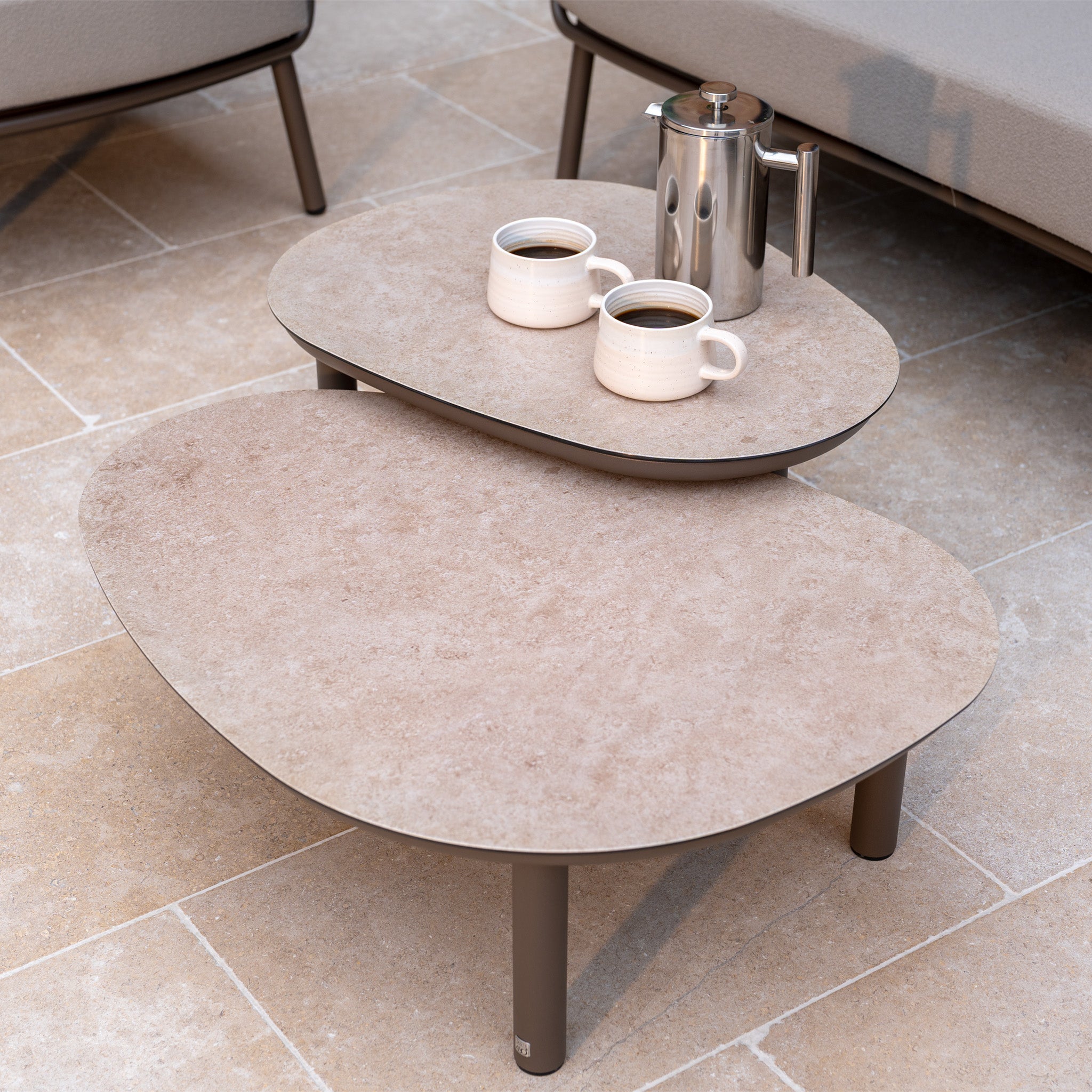 Two coffee cups and a metal coffee pot on nested stone-top tables with beige tile flooring, perfectly complementing your Capri 3 Seat Sofa Set in Stone Grey.
