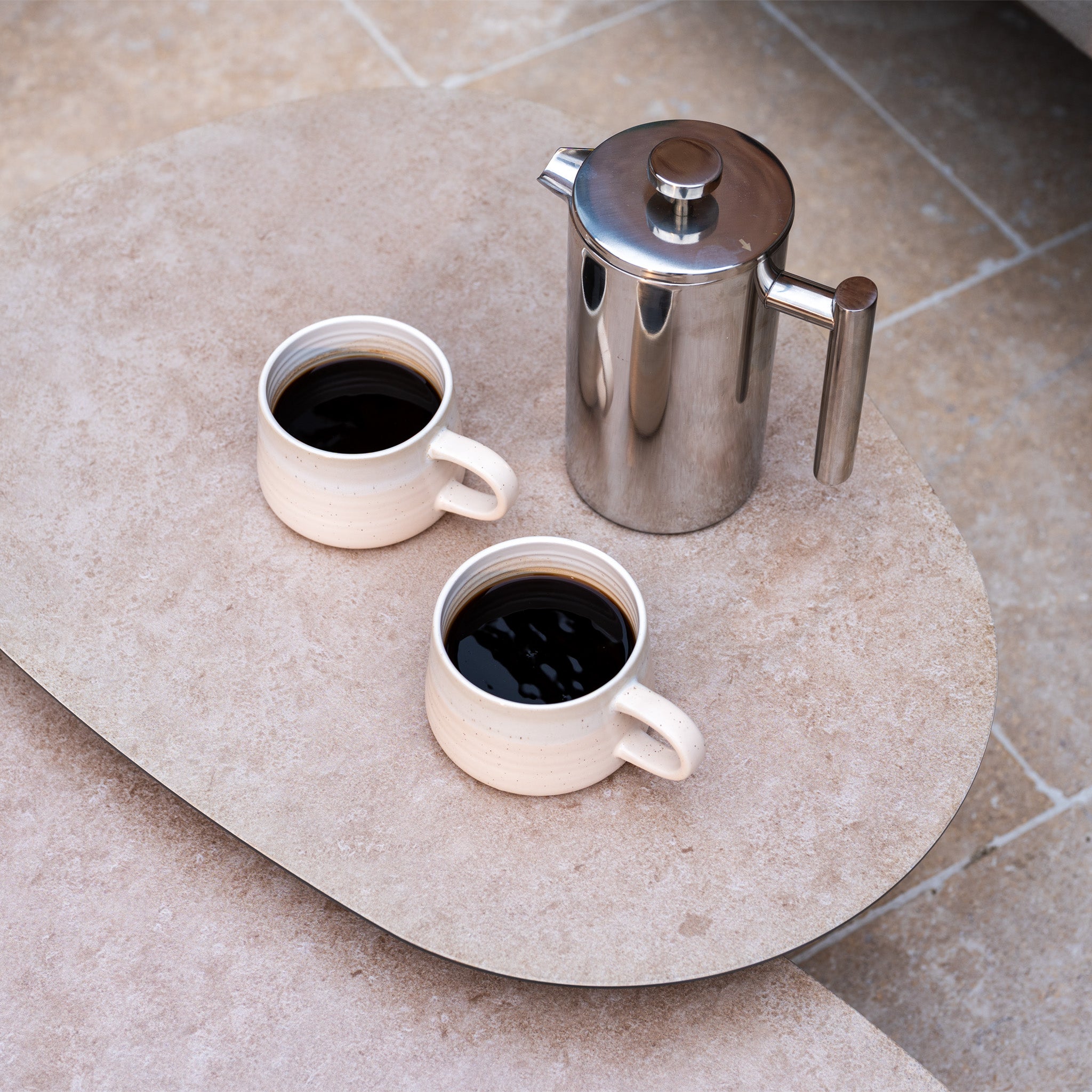 Two white mugs with black coffee and a stainless steel French press rest on a stone table, perfectly complementing the Capri 3 Seat Sofa Set in Stone Grey for a cozy outdoor experience.