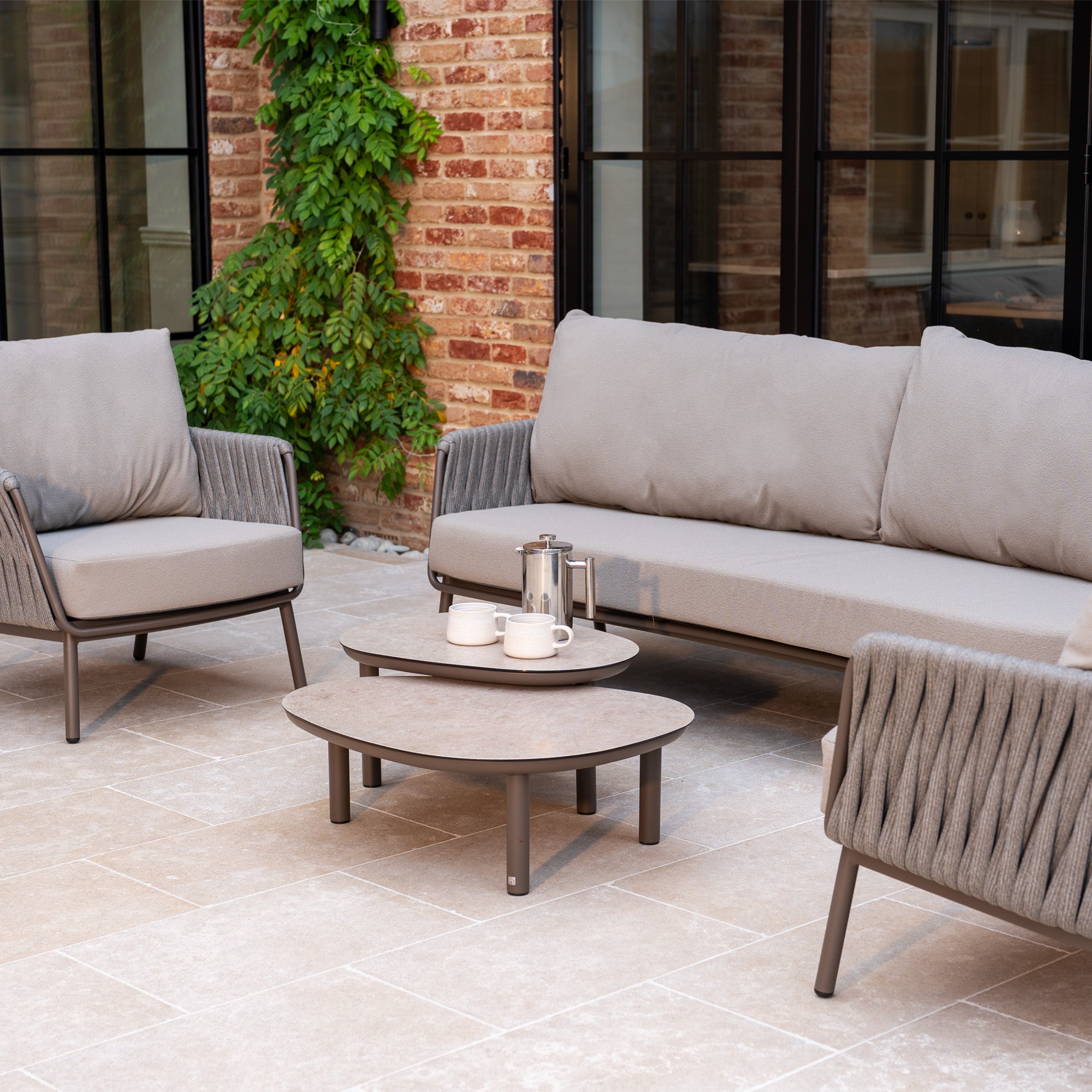 Modern patio with a Capri 3 Seat Sofa Set in Stone Grey and a coffee table, set against a brick wall with green vines for a stylish finish.