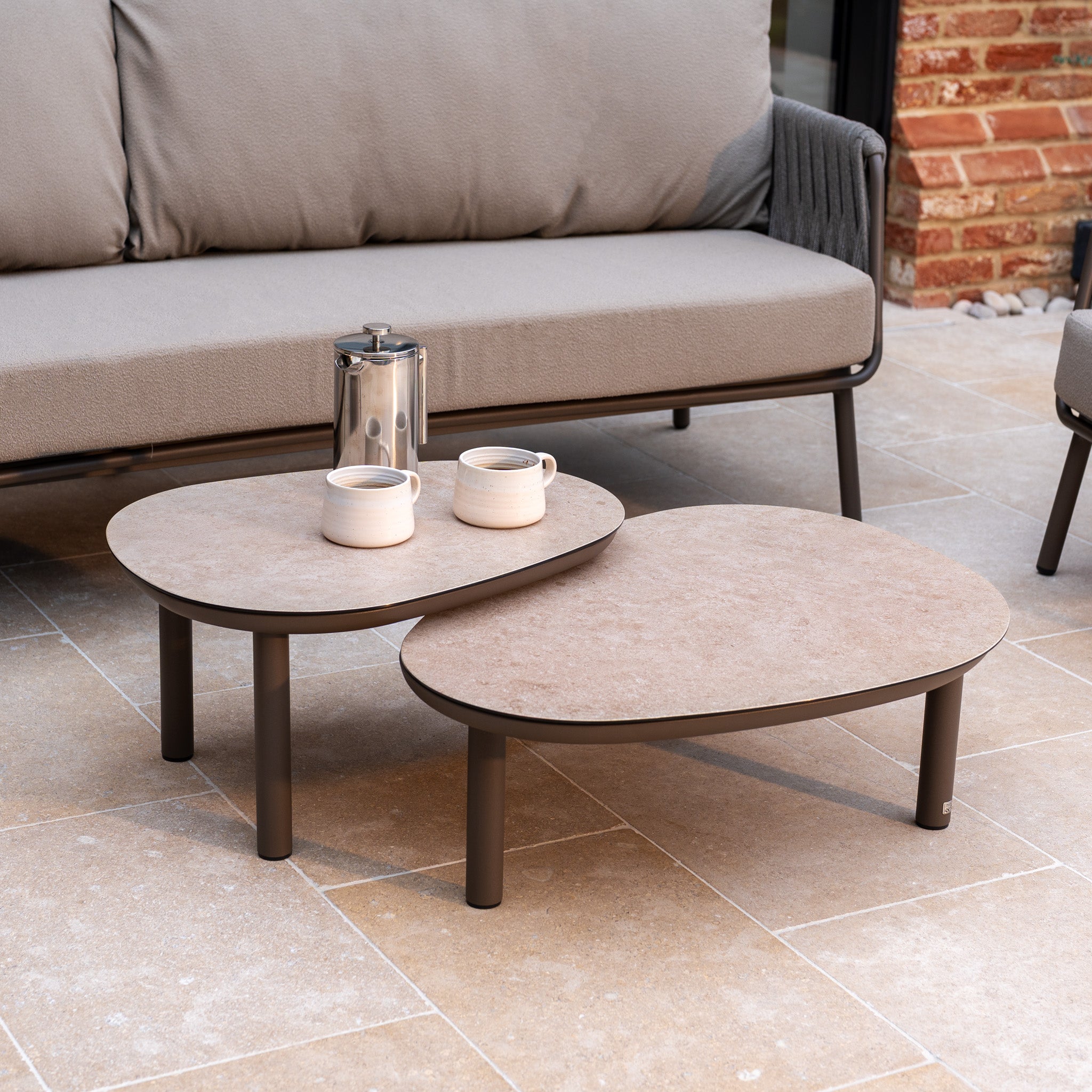 Two oval coffee tables with a coffee pot and two mugs are placed before a Capri 3 Seat Sofa Set in Stone Grey on a tiled patio, creating an inviting garden furniture setup.