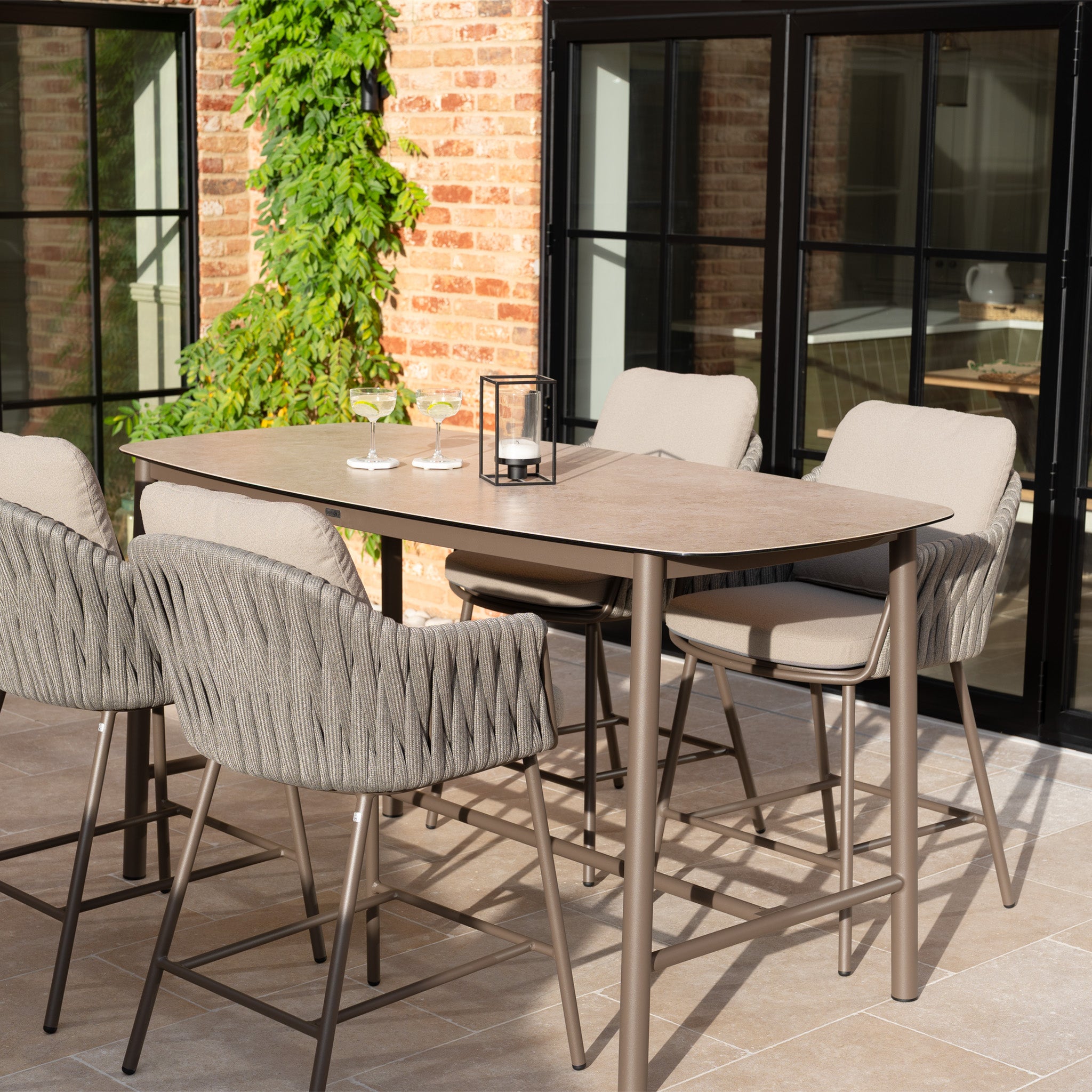 The Capri 4 Seat High Rectangular Dining Set in Stone Grey features cushioned chairs, perfect for enjoying drinks on a patio beside brick and glass walls.
