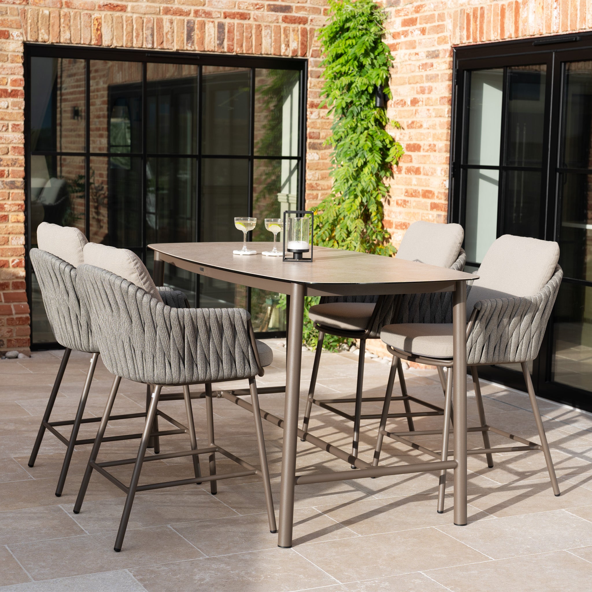 The Capri 4 Seat High Rectangular Dining Set in Stone Grey includes four cushioned chairs and a modern table, perfect for elevating your outdoor space with contemporary style near brick walls and windows.